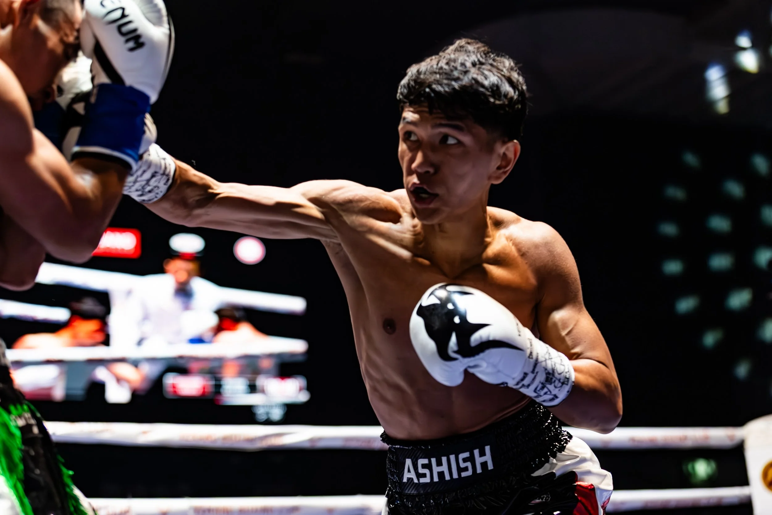 A shirtless male boxer throwing a punch in a boxing ring, wearing boxing gloves and shorts labeled 'ASHISH', with a digital screen in the background showing a referee and other fighters.