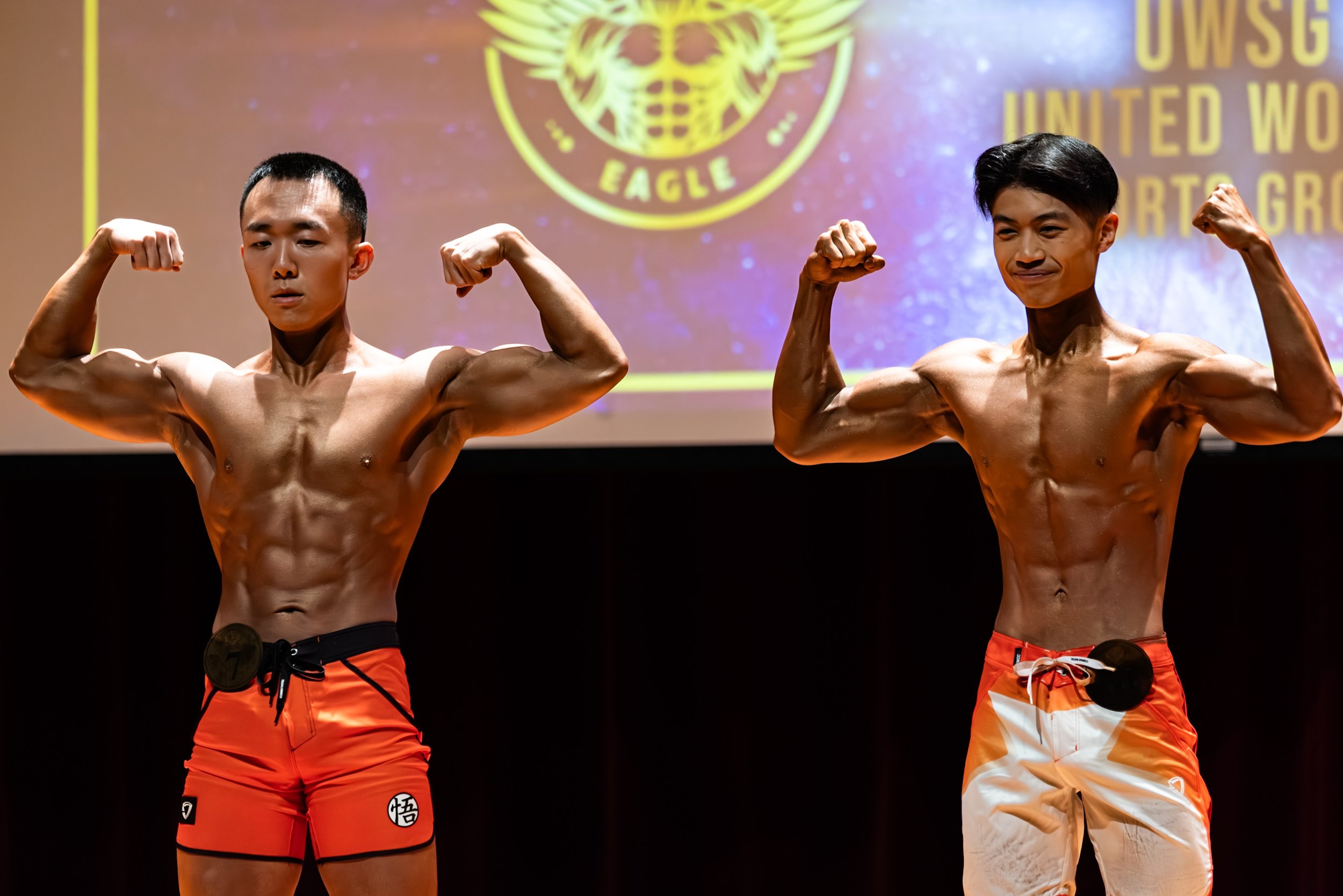 Two shirtless male bodybuilders flexing their arm muscles in a competition setting, with a background display of a logo and text.