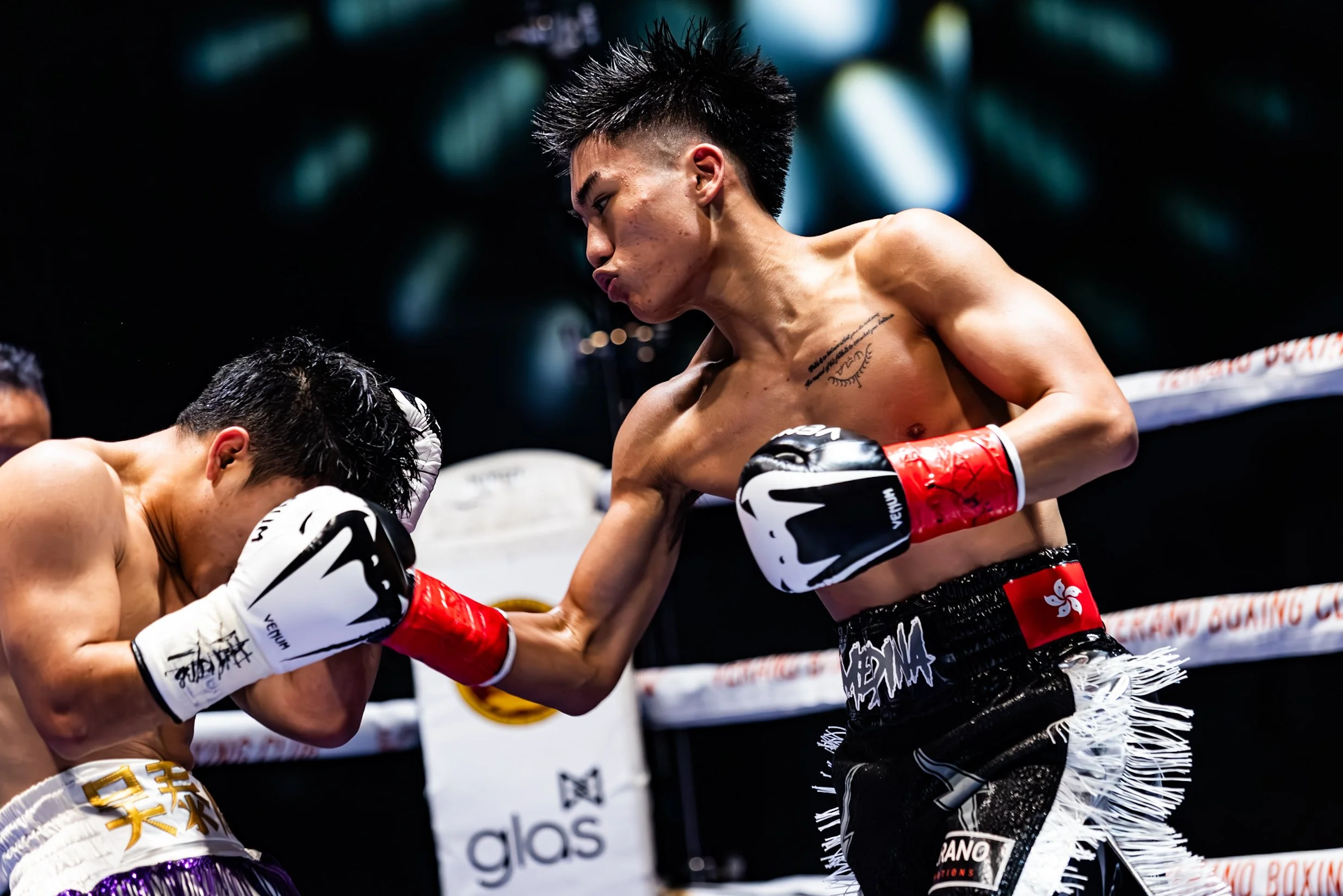 Two male Muay Thai fighters engaged in a fight inside a ring. The fighter on the left is covering his face with his gloves, while the fighter on the right is throwing a punch. Both are wearing boxing gloves and shorts.