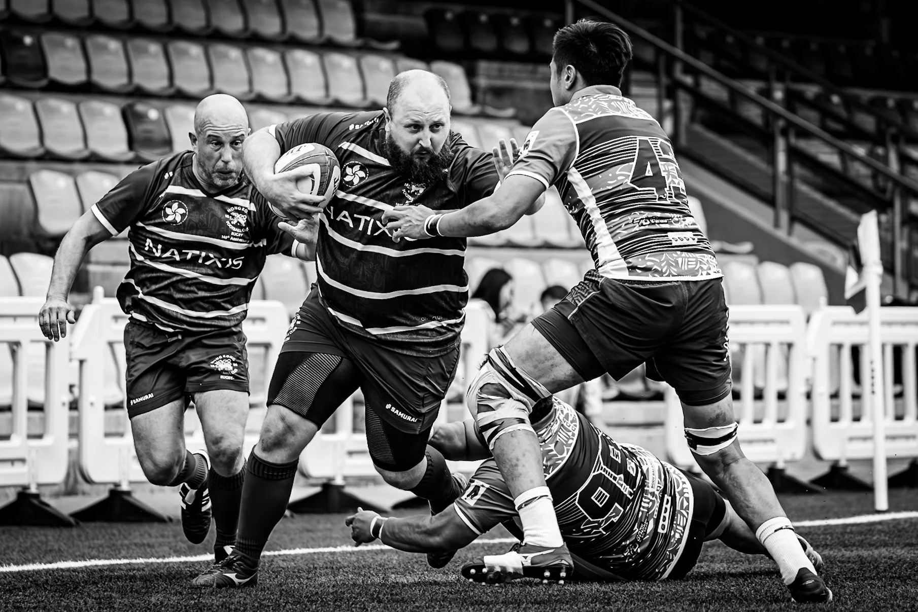 Black and white photo of rugby players in action, with one player holding a rugby ball while being tackled by another player on the field.