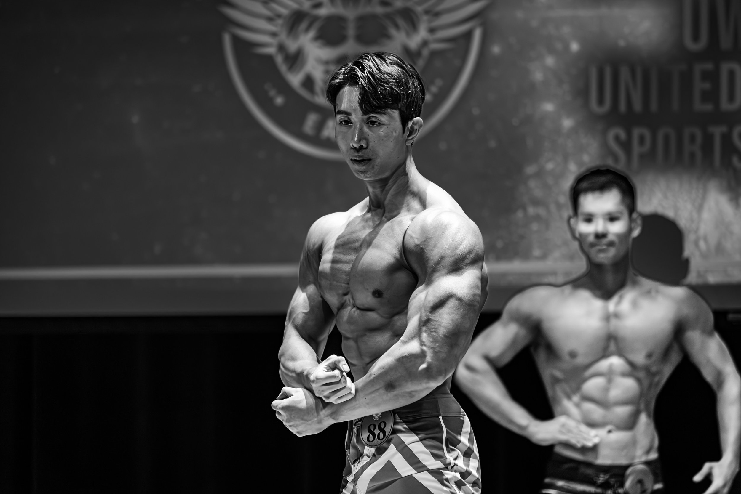 Black and white photo of two male bodybuilders on stage at a competition, one in the foreground flexing his muscles and the other in the background with his hands on his hips.