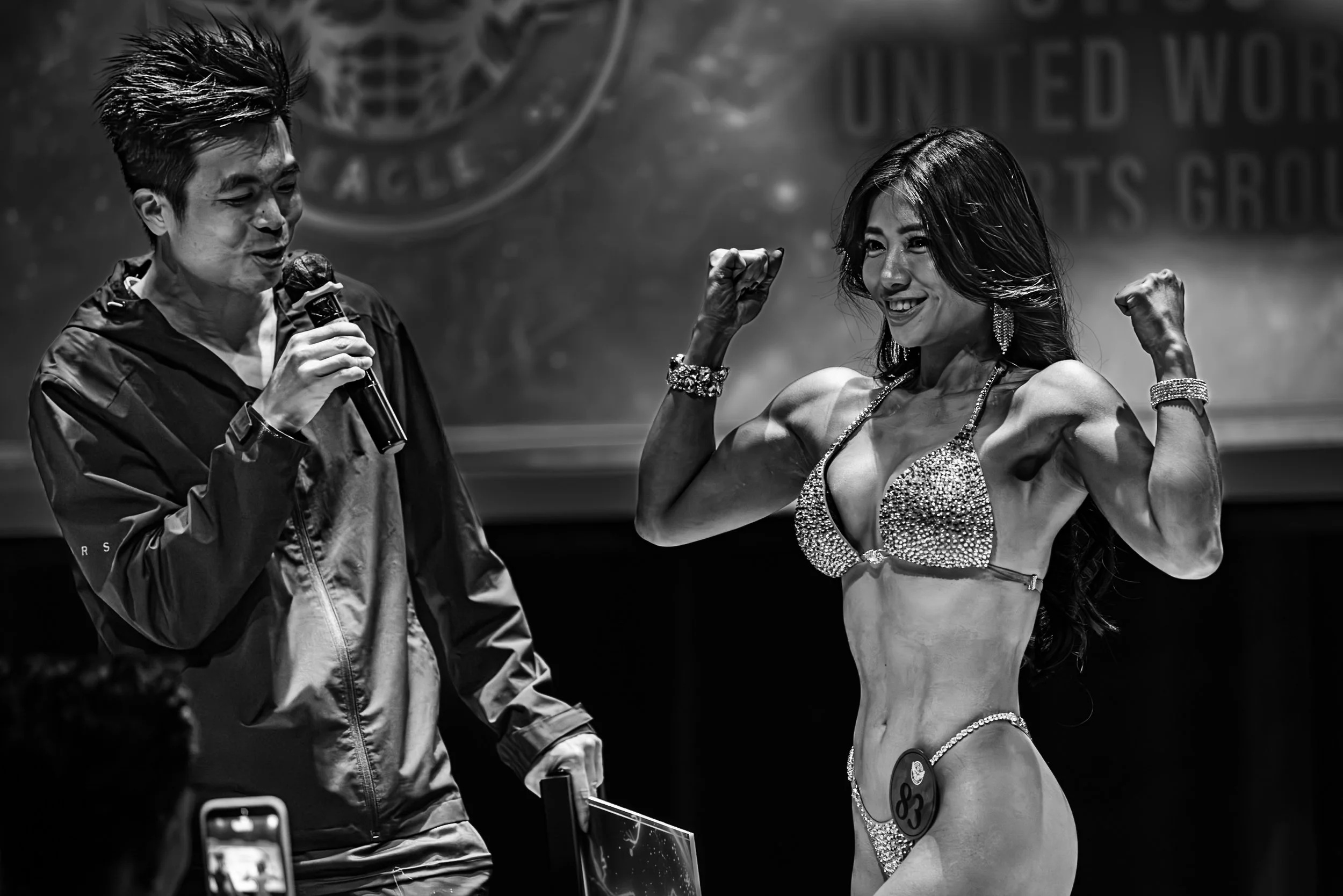 A female bodybuilder in a bikini flexing her muscles on stage, smiling and showing off her physique, while an interviewer holds a microphone nearby.