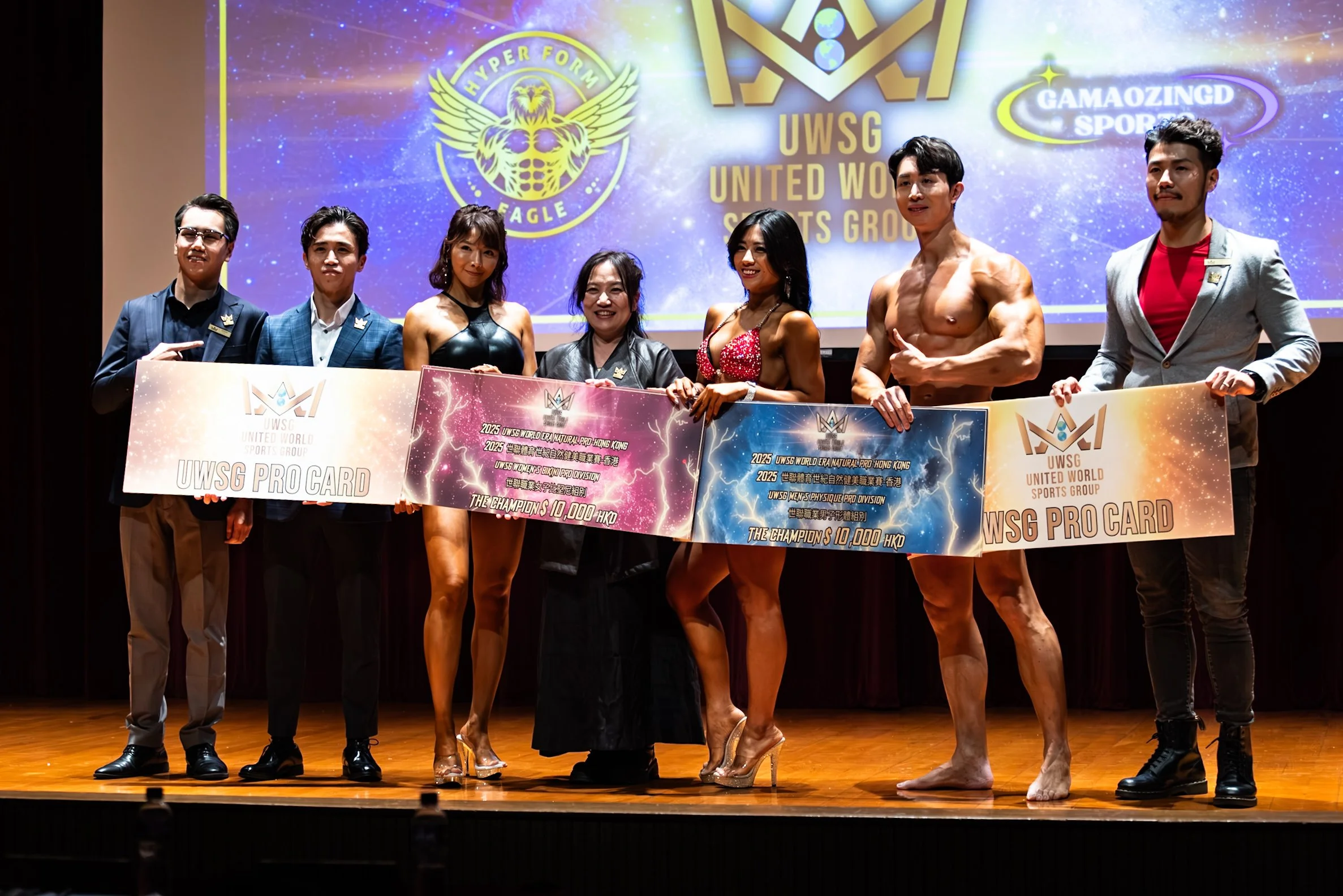 Group of seven people standing on stage holding large presentation boards and smiling at the camera, with a bright colorful backdrop displaying various logos and text in the background.
