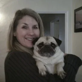 Woman smiling and holding a small pug dog indoors.