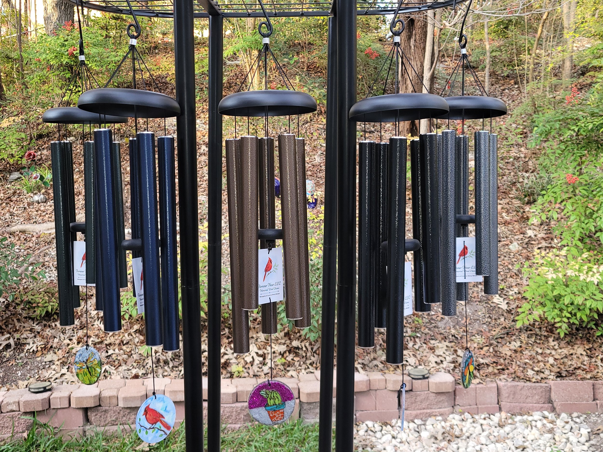 A set of black wind chimes with colorful handcrafted decoration hanging underneath them, positioned outdoors on a garden with trees and shrubs.