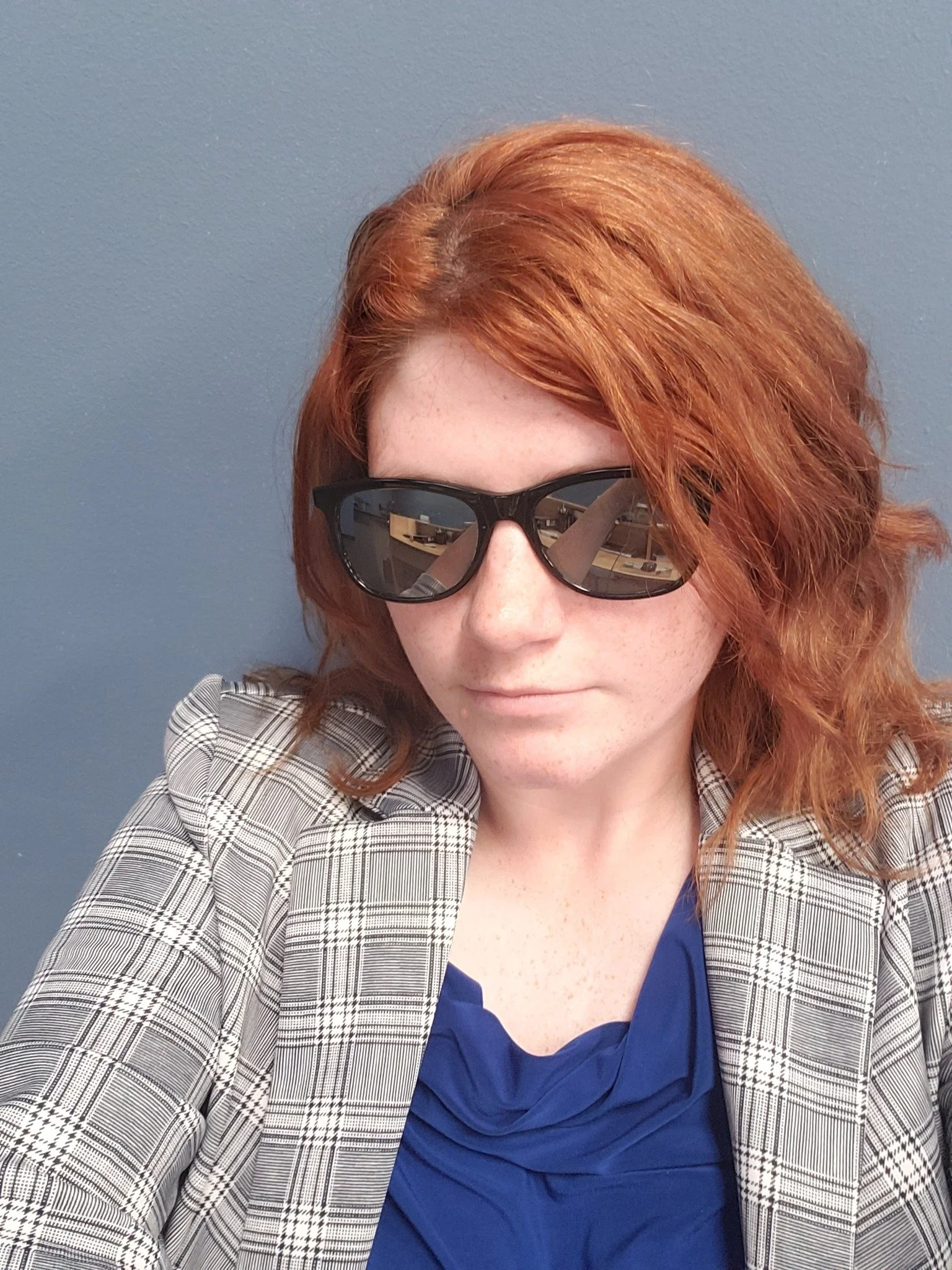 A woman with red hair wearing sunglasses, a blue top, and a plaid blazer standing against a blue wall.