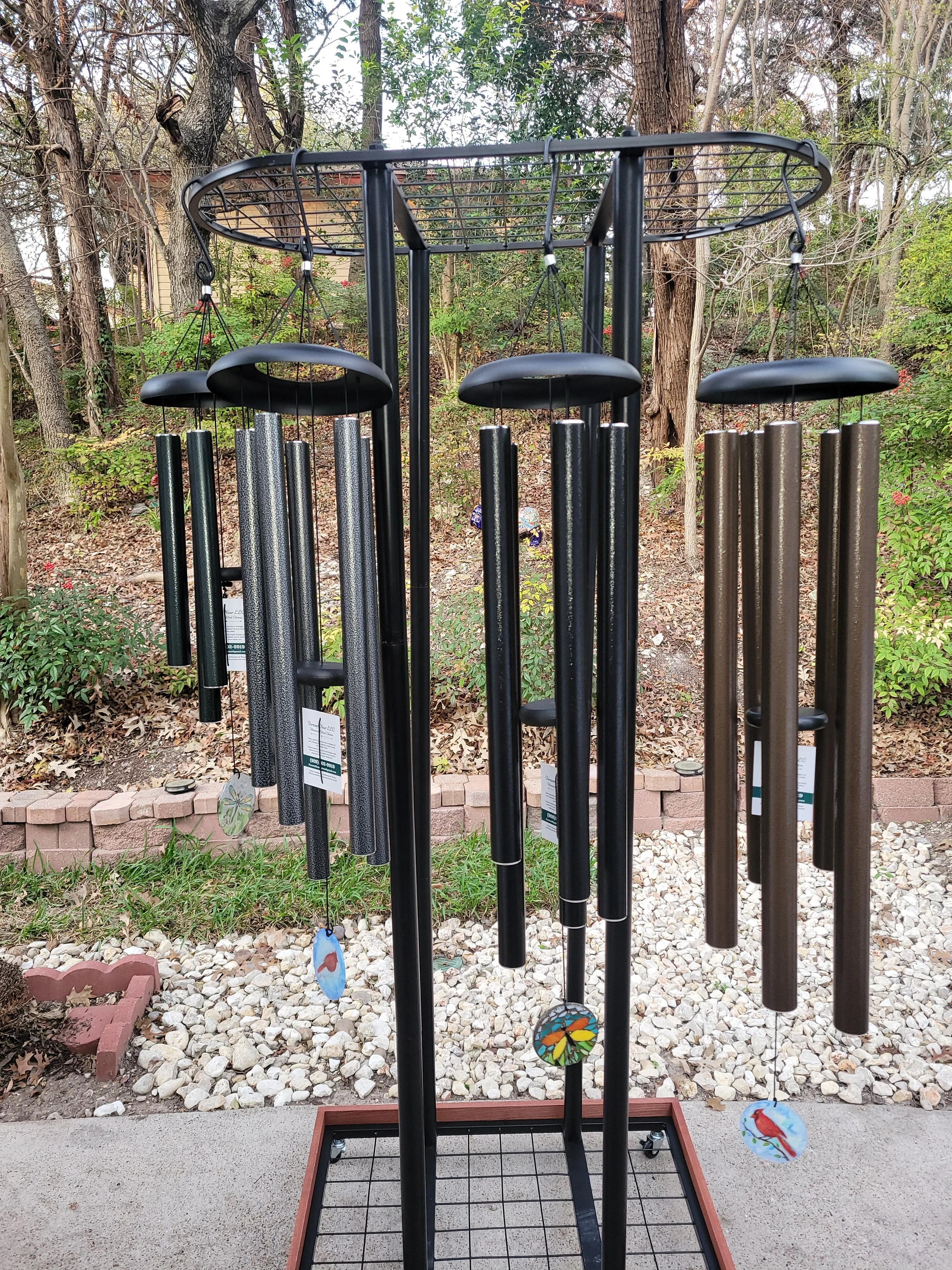 A wind chime stand with multiple black and metallic chimes hanging from a black metal frame outdoors in a garden with trees, plants, and a brick border.