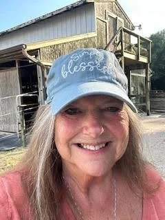 Smiling woman wearing a light blue cap with 'Some folks are just blessed' written on it, standing outdoors in front of a rustic wooden barn and shed.