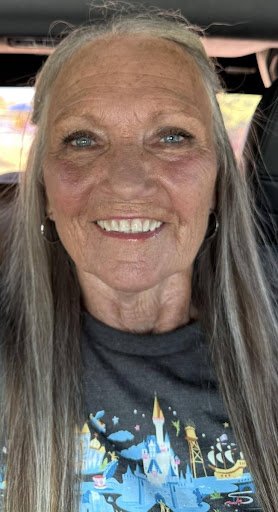 Close-up of an elderly woman with gray hair, blue eyes, and a big smile, inside a vehicle, wearing a dark t-shirt with a Disney castle design.