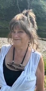 Young woman with messy hair wearing a light blue top and sunglasses hanging around her neck, outdoors near water with trees in the background.