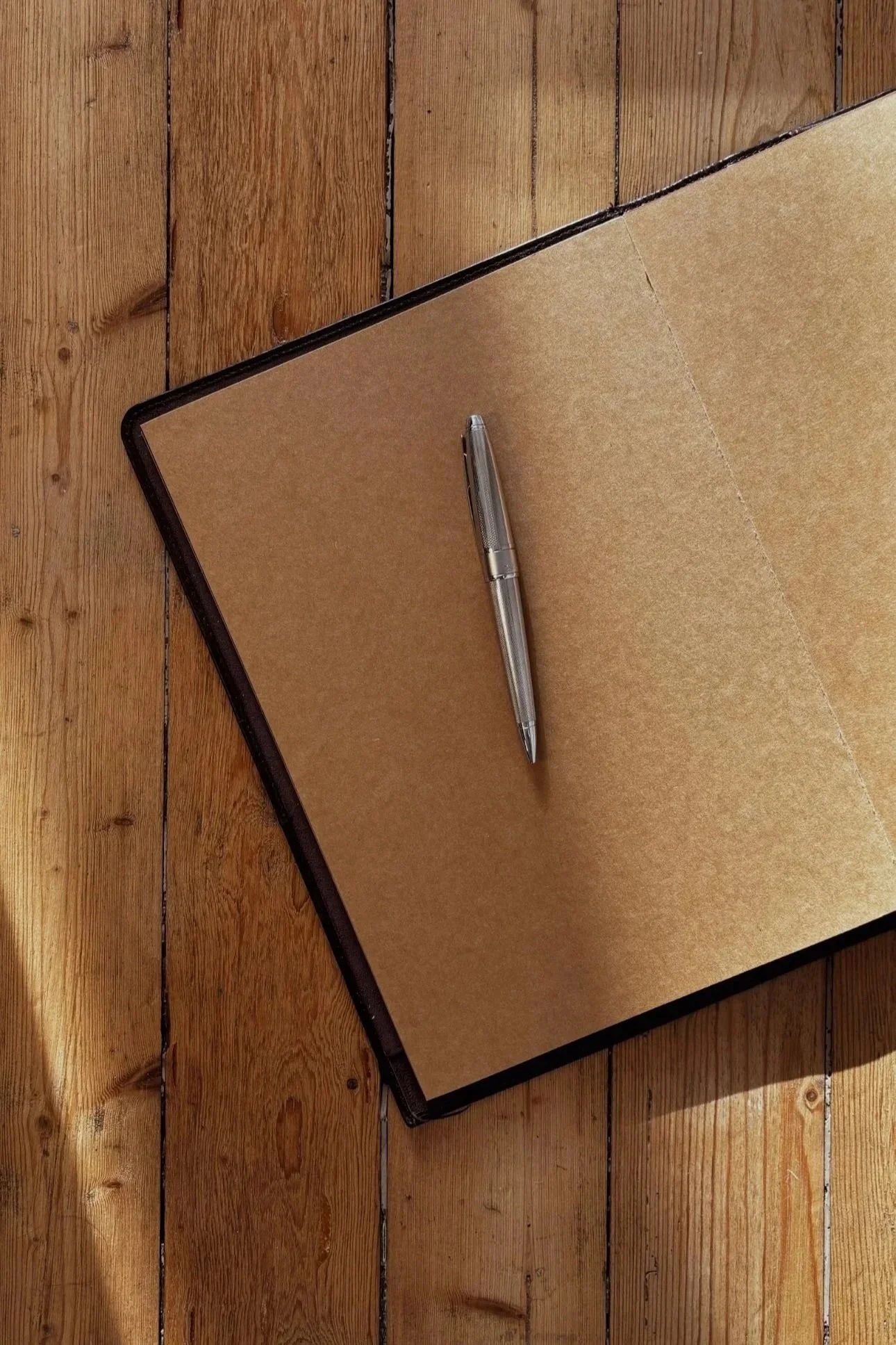 Un cahier ouvert avec une couverture en carton et une balle stylos argentée posé dessus, sur un plancher en bois.