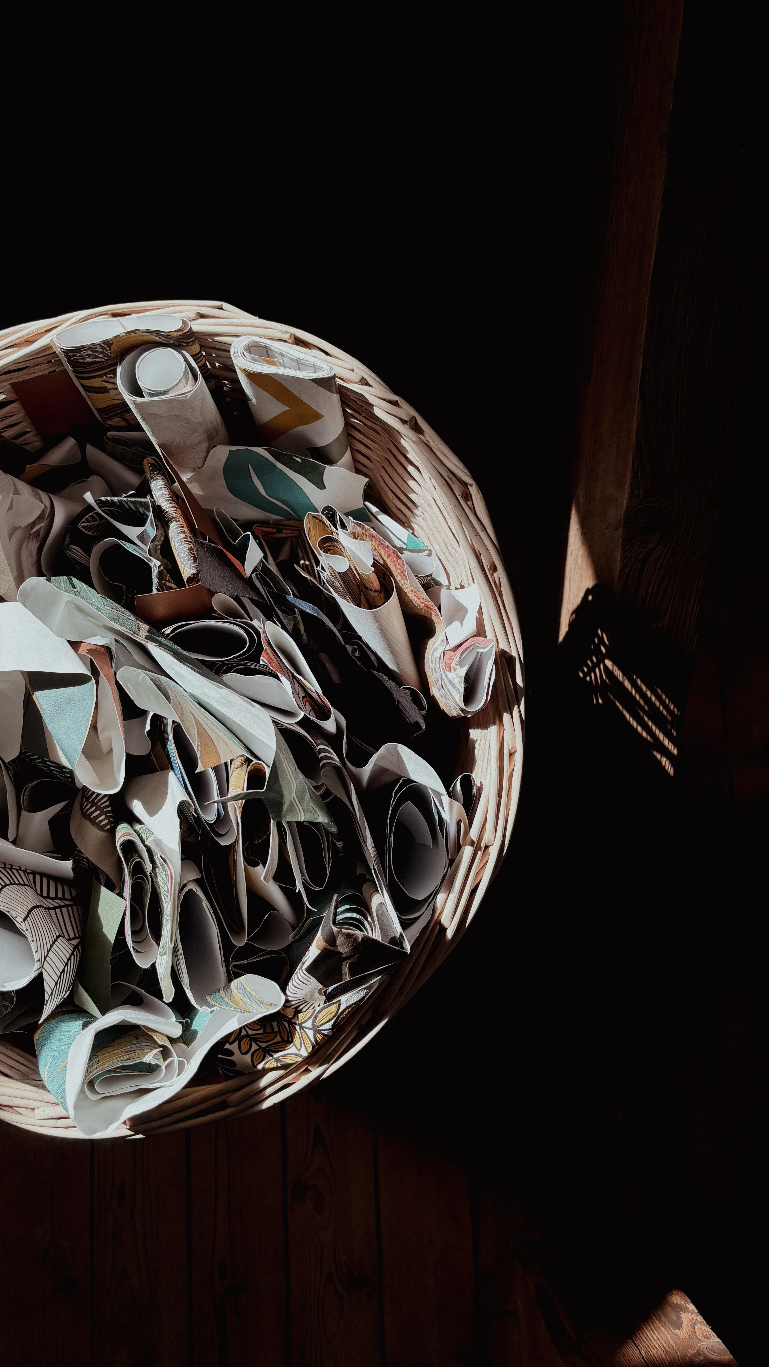 A wicker basket filled with crumpled, patterned paper or fabric scraps, sitting on a wooden surface with a shadow cast from sunlight.
