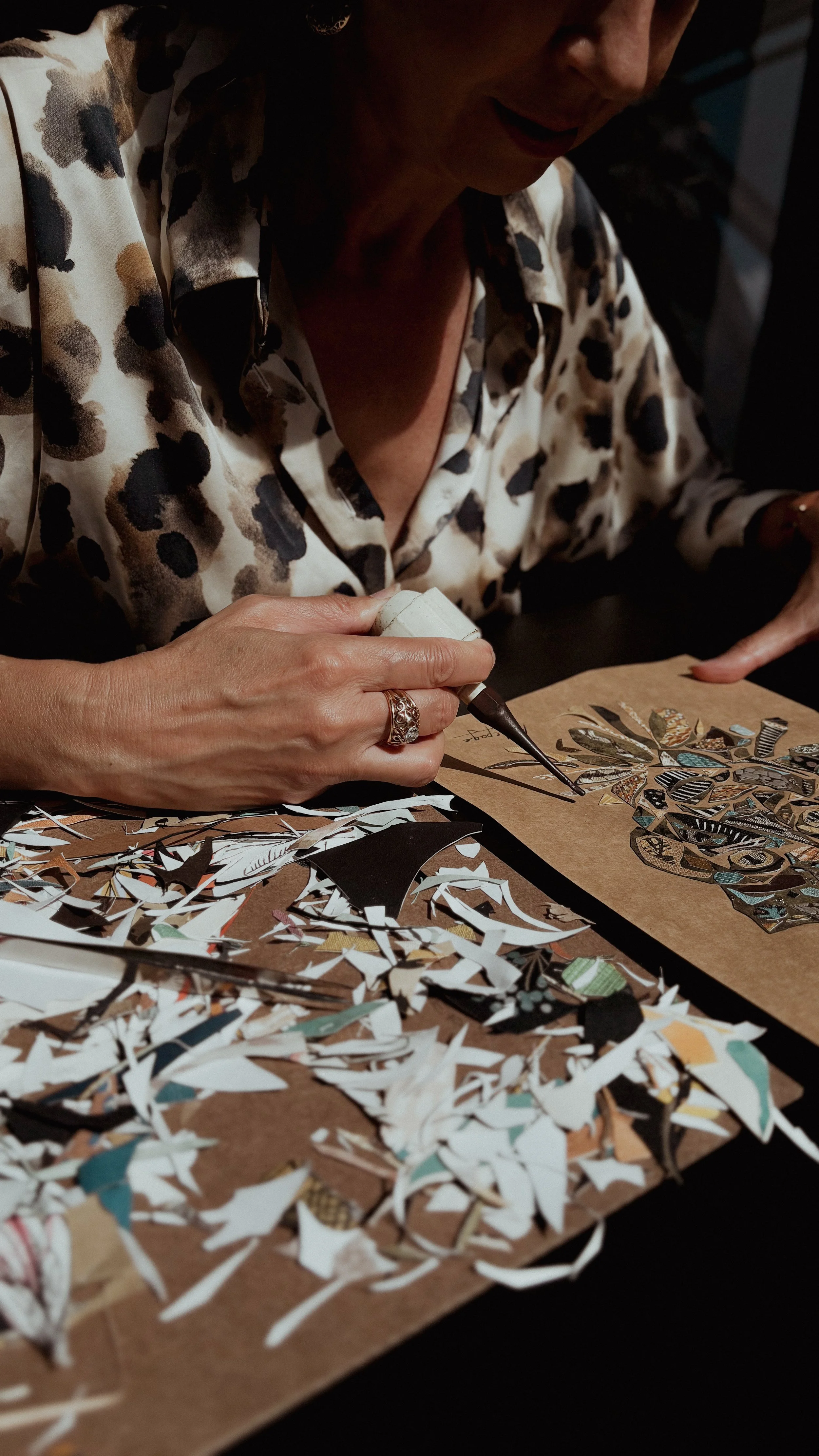A woman wearing a leopard print blouse is creating artwork with paper pieces, using a glue applicator, surrounded by torn paper scraps on a table.