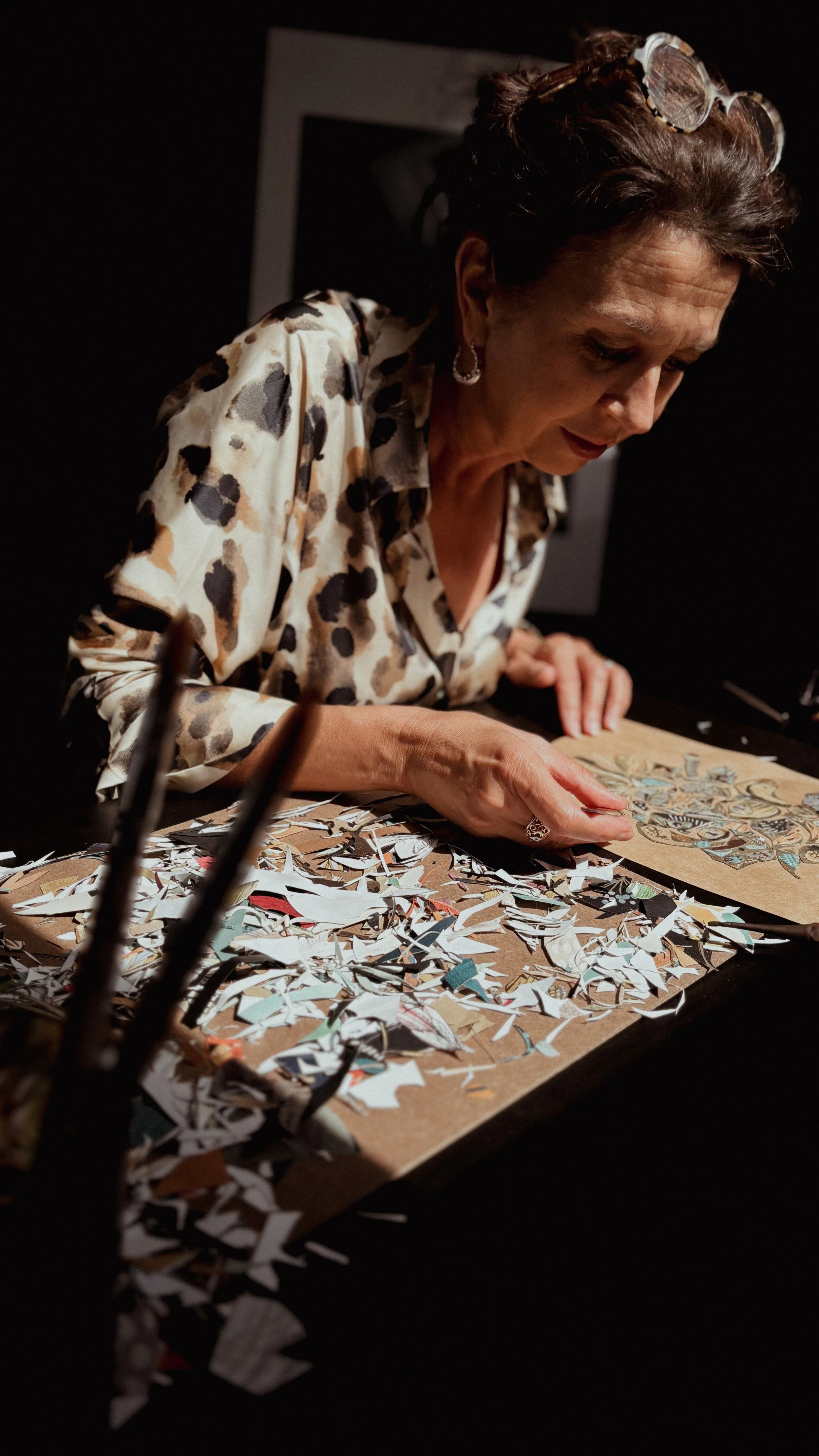 A woman with glasses on her head, wearing a leopard print blouse, is working on a collage of torn paper pieces on a wooden board in a dimly lit space.
