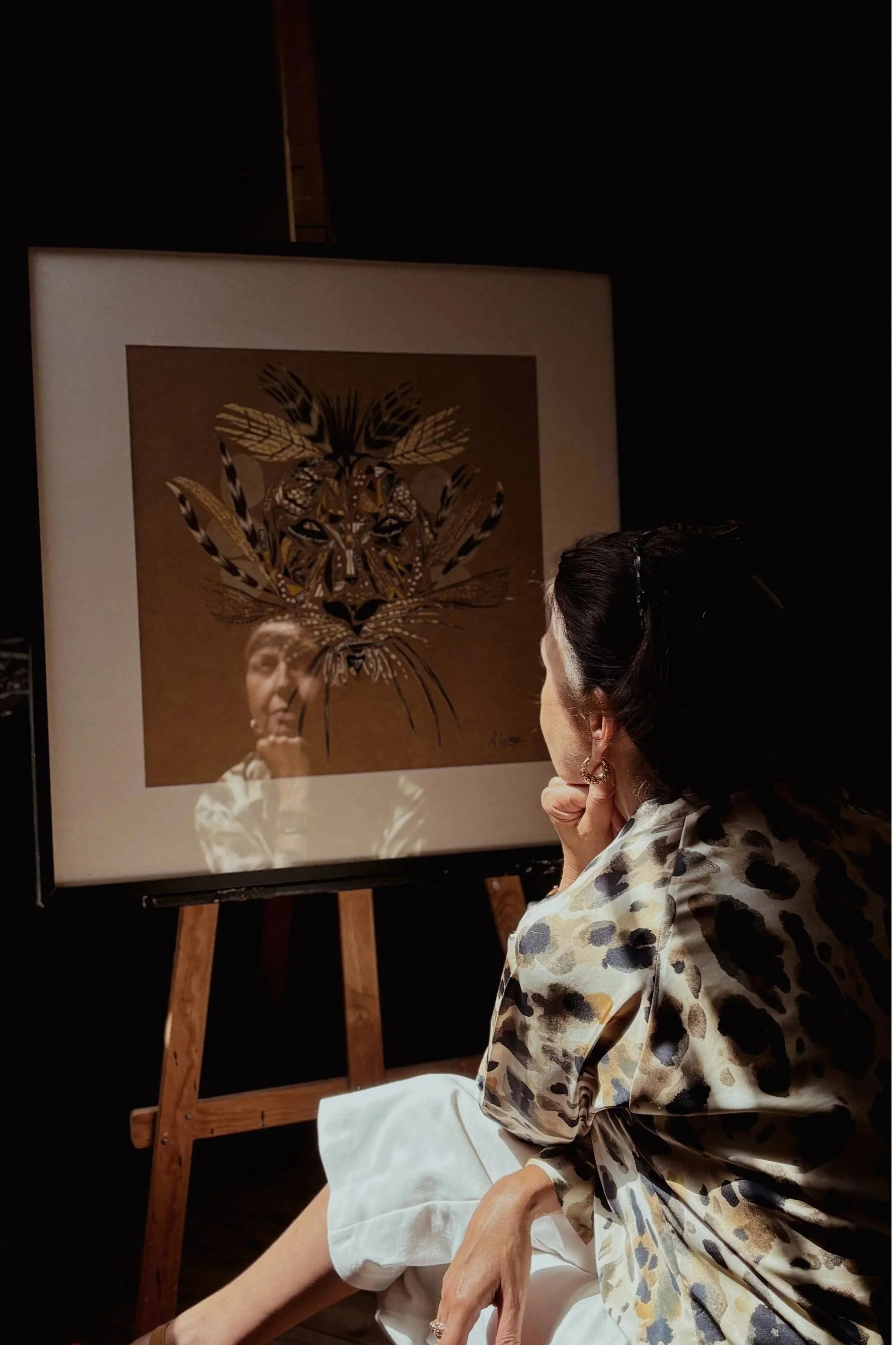 Aline Lepage sitting and observing an artwork of a tiger's face, which is a collage of various feather patterns, displayed on an easel.