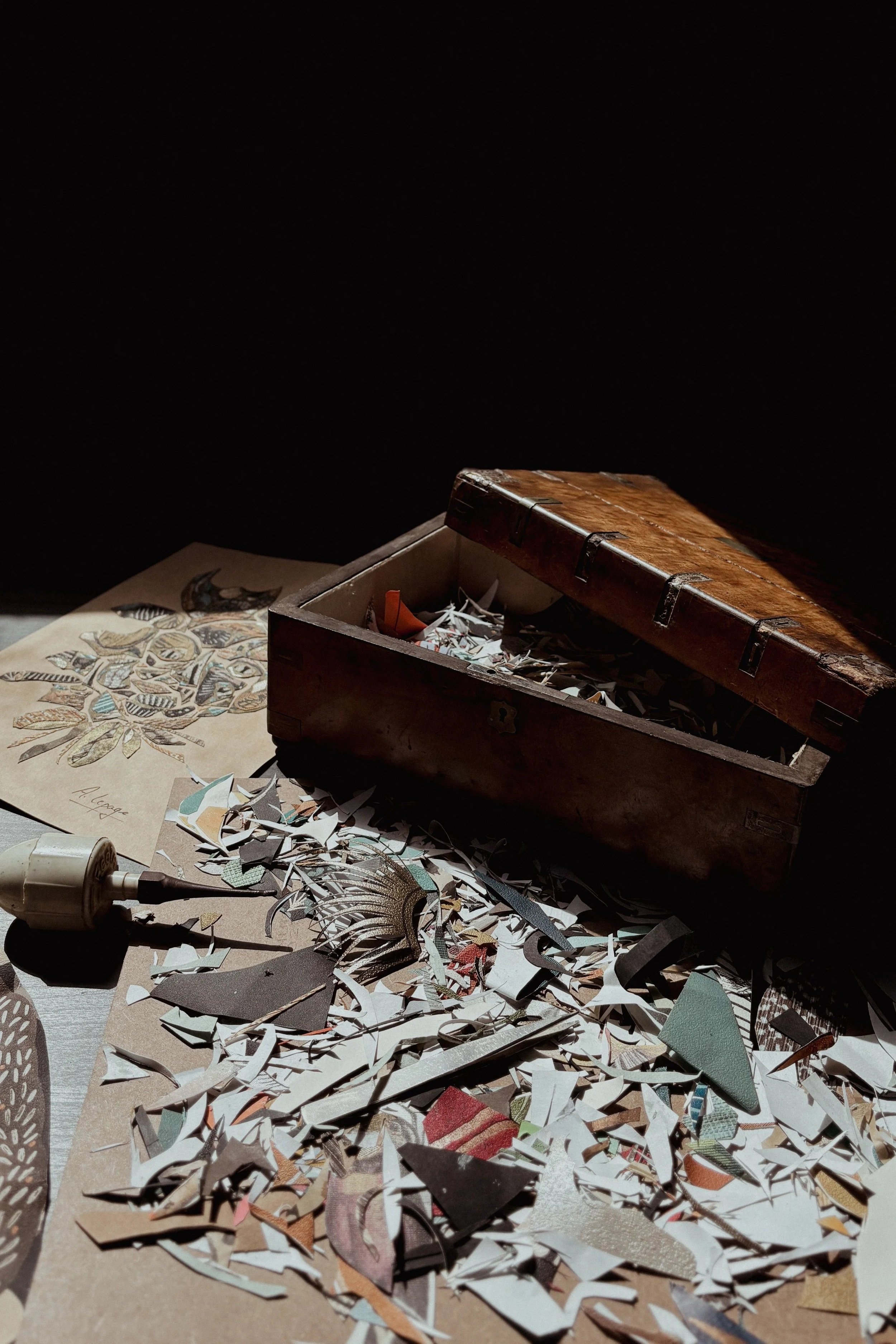 A wooden box surrounded by scattered paper cutouts of various colors and patterns, with an illustration of botanical elements beside it and a tool with a pointed tip on the table.