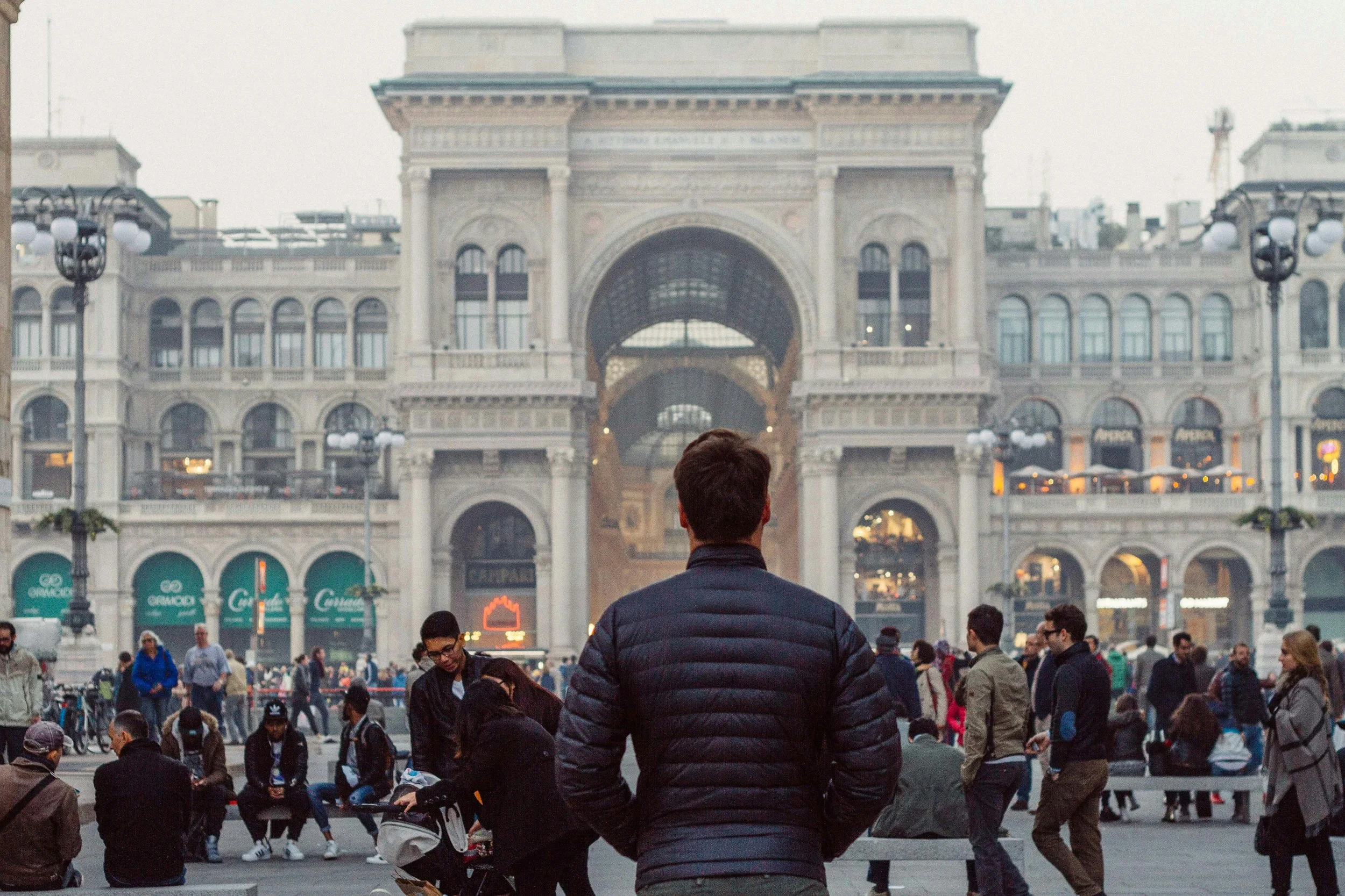 Una piazza affollata con una persona in primo piano che guarda verso un grande edificio con archi e finestre, in un ambiente urbano con molte persone che camminano e si riuniscono.