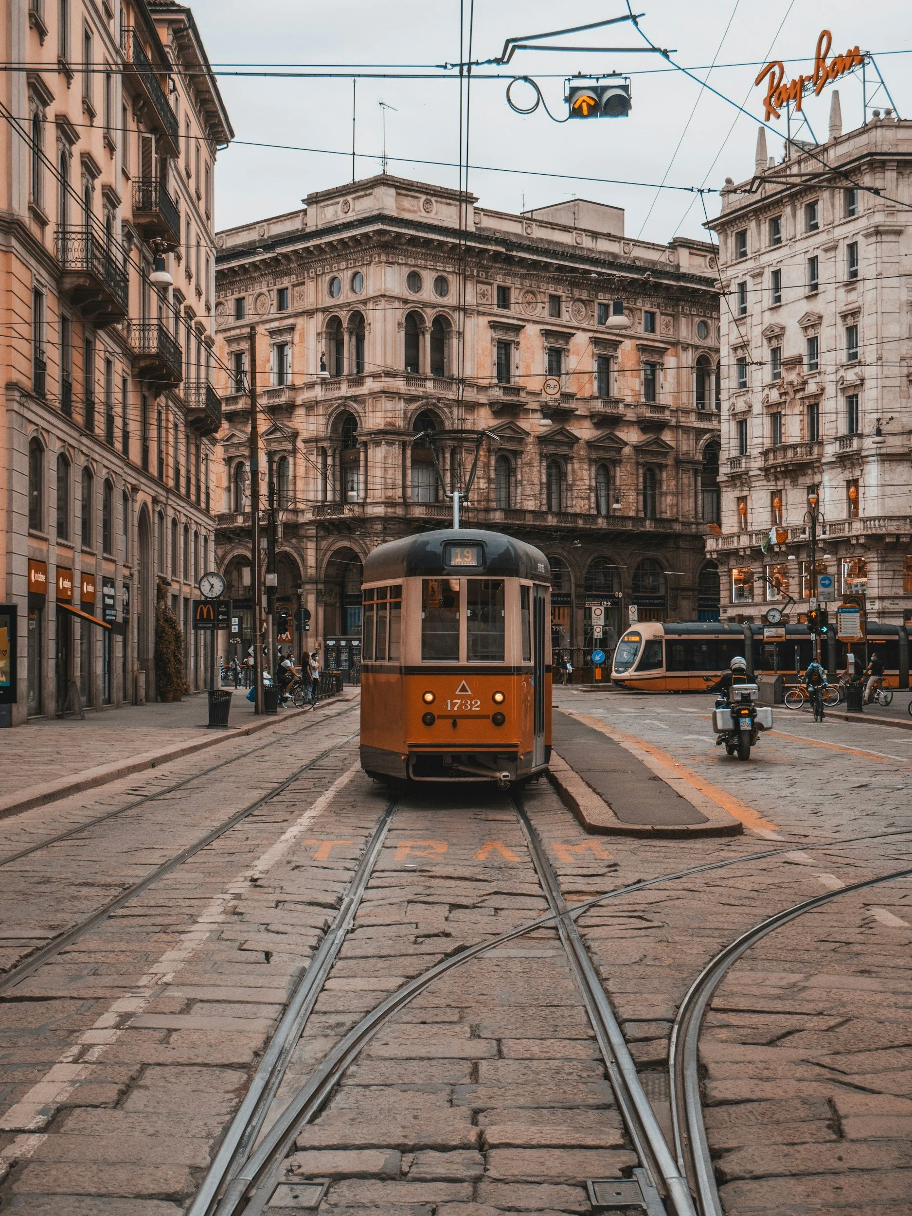 Una strada urbana con tram arancione che si avvicina, edifici storici e un cielo grigio sopra, con persone in bicicletta e motorini ai lati.