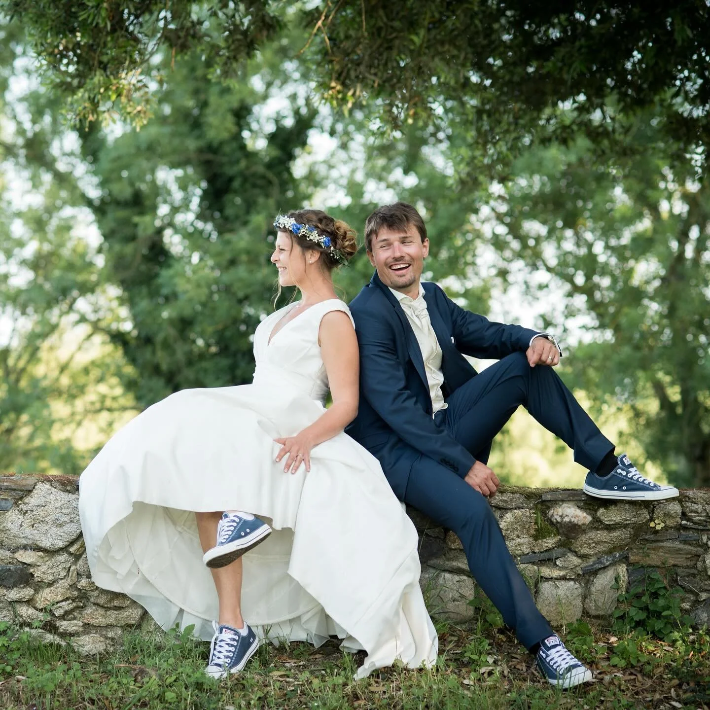 Un couple de mariés portant des chaussures de sport, assis sur une murette en pierre dans un parc verdoyant
