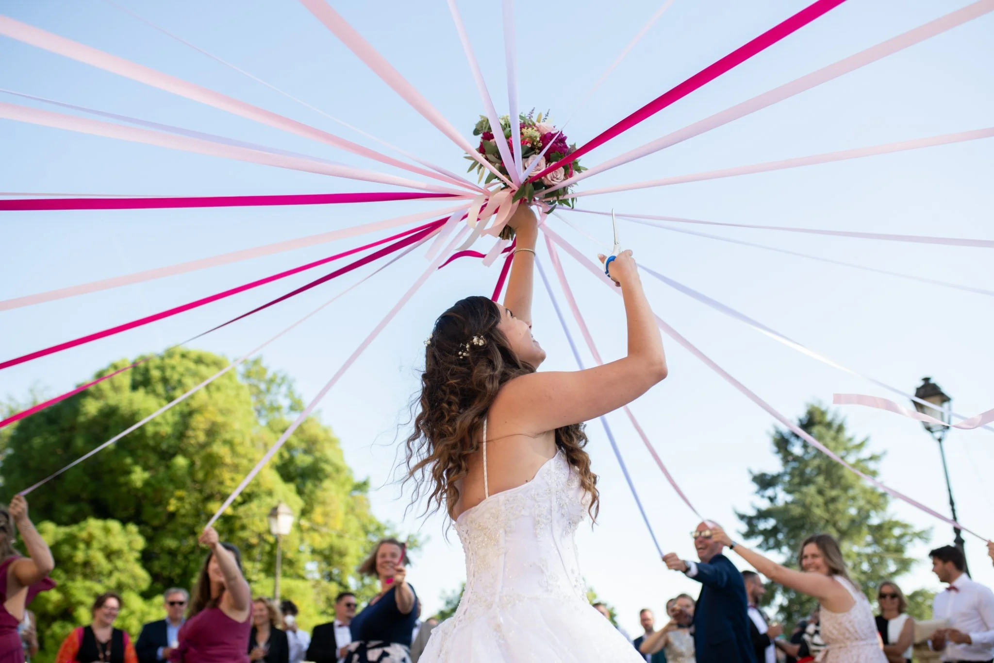 Une mariée en robe blanche lors d'une fête de mariage, levant un bouquet de fleurs avec des rubans colorés lors d'une cérémonie en plein air, entourée de membres du cortège et d'invités sous un ciel clair.