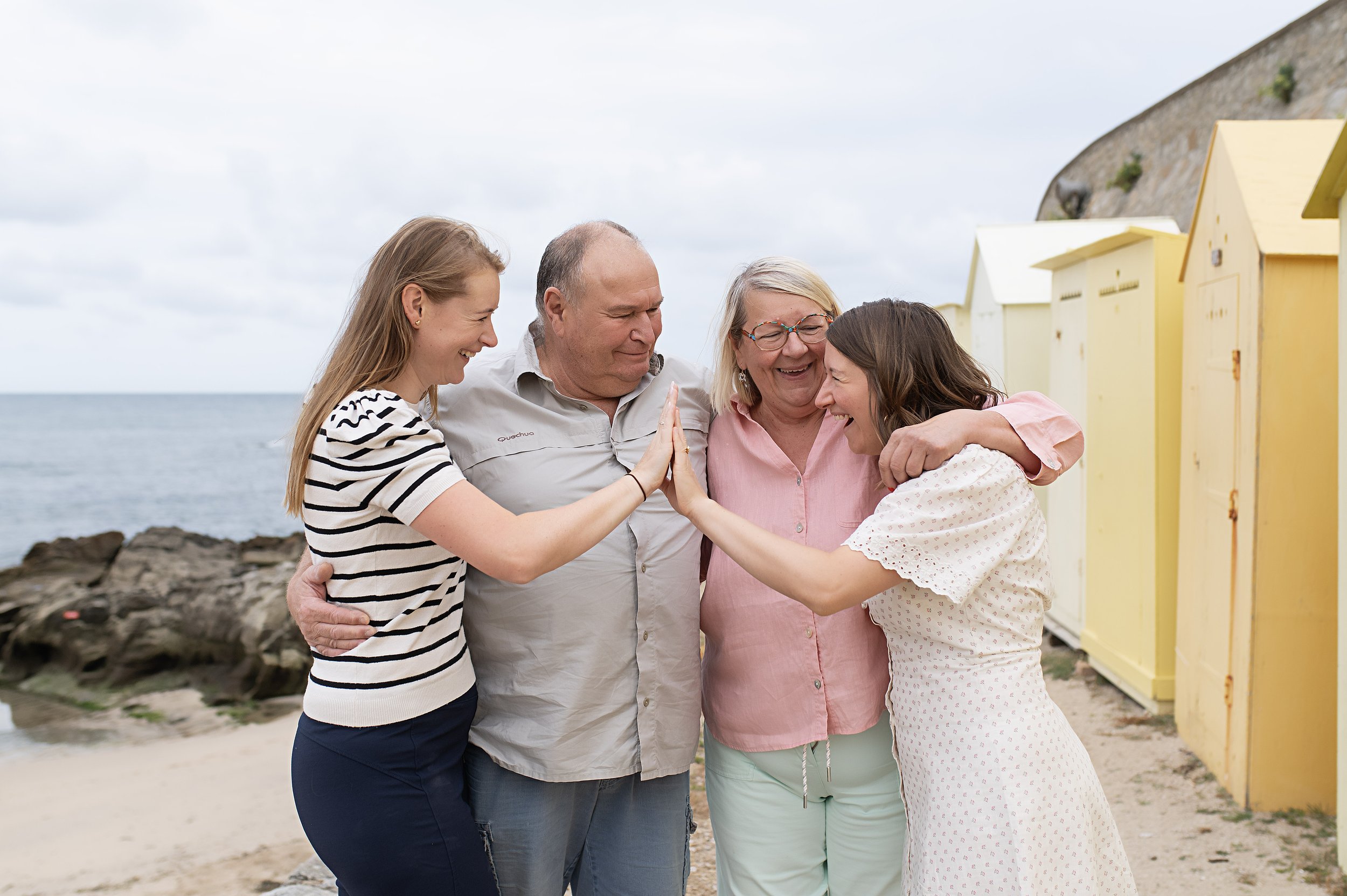 Quatre personnes souriantes en famille en train de se faire un high-five sur la plage, avec des cabines de plage jaunes en arrière-plan.