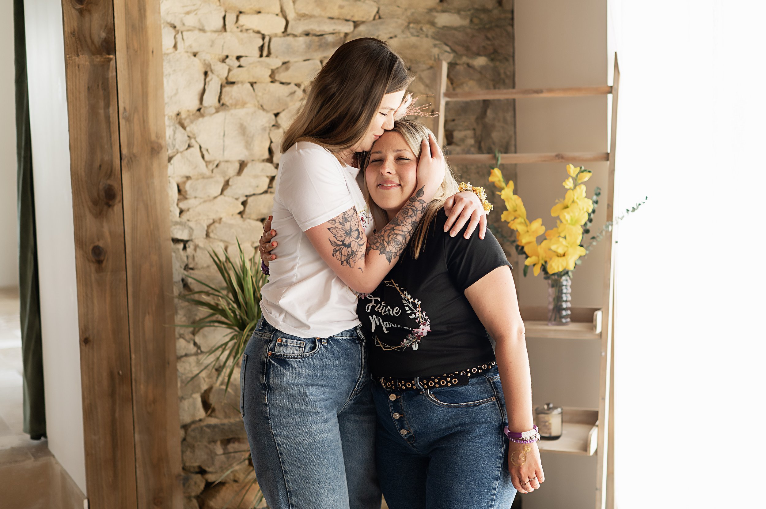Deux femmes s'étreignant avec émotion dans une pièce décorée de plantes et de fleurs. La femme à gauche porte un t-shirt blanc, et la femme à droite porte un t-shirt noir avec une inscription, avec un fond en pierre et en bois.