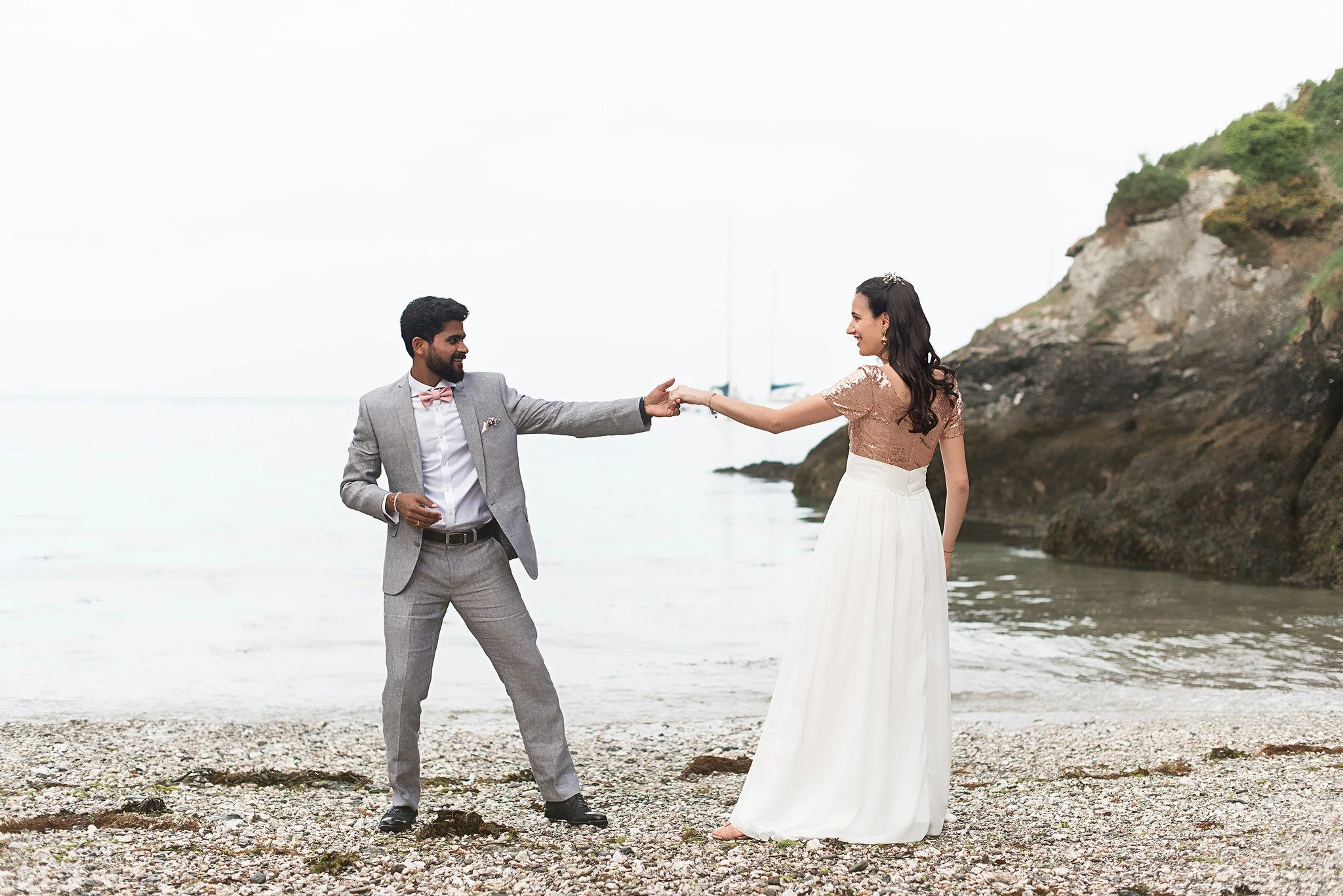 Un couple en vêtements de mariage s'amuse sur une plage rocheuse avec la mer en arrière-plan, le homme tend la main à la femme.