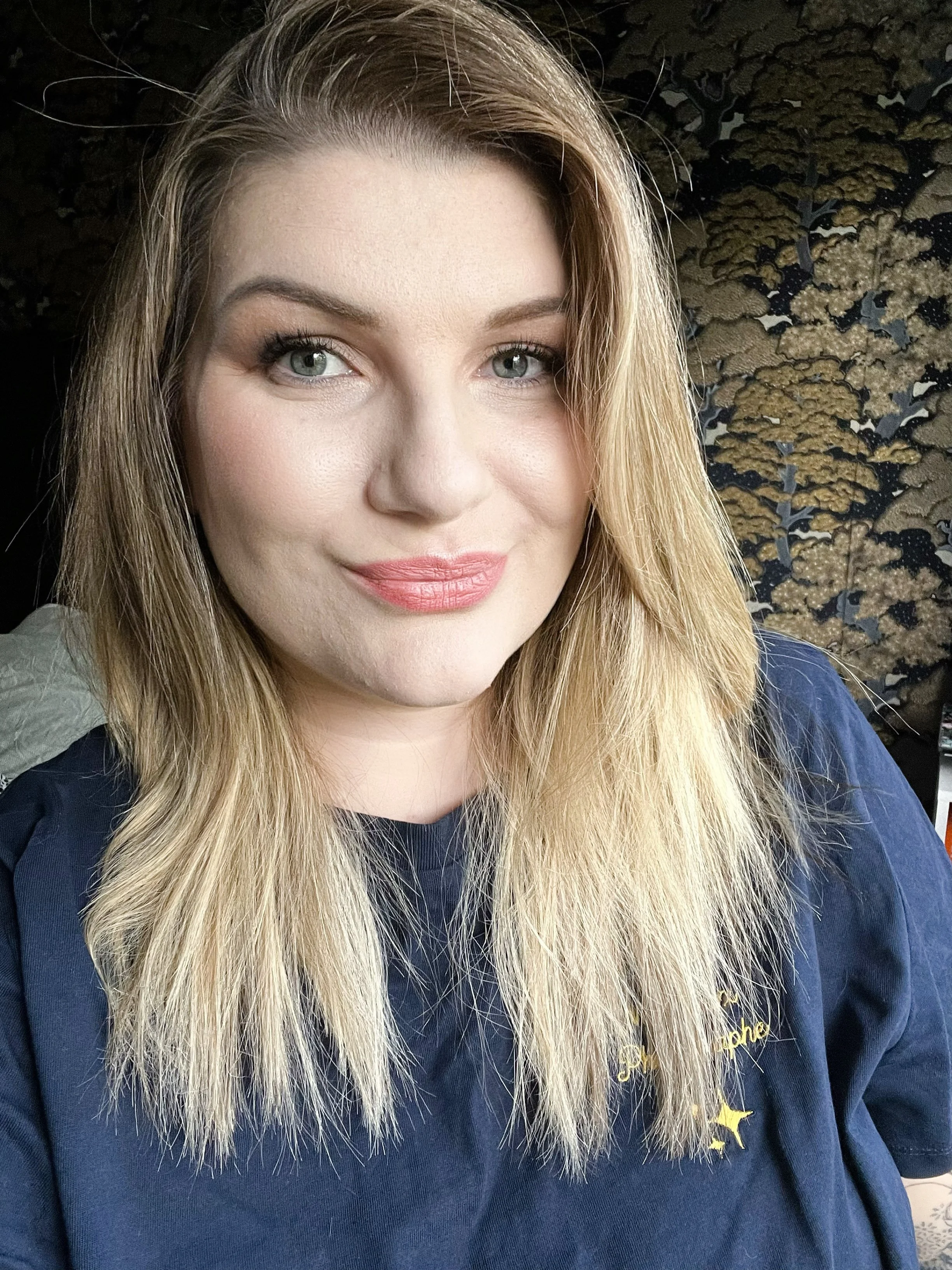 Une femme avec des cheveux blonds et longs, porte un t-shirt bleu, sourit à la caméra, en arrière-plan un mur avec un motif de feuilles ou d'arbres dorés et noirs.