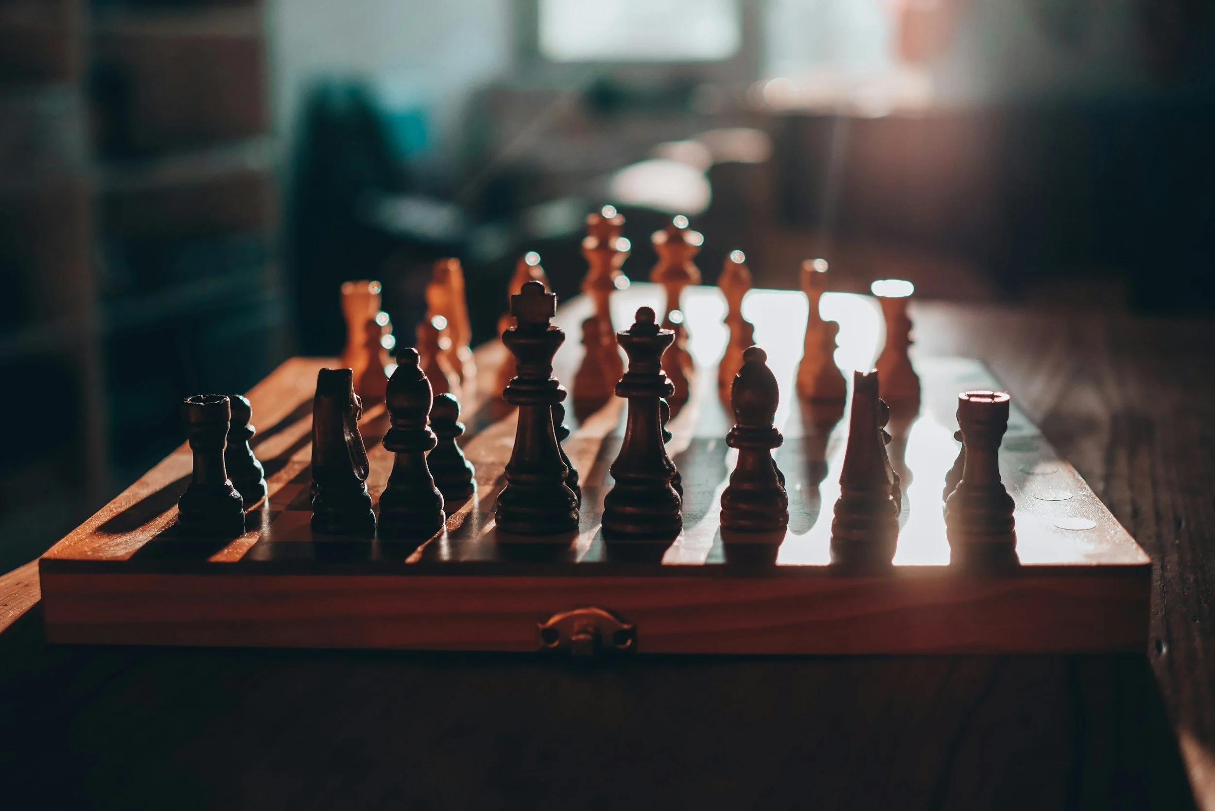 A wooden chessboard set up for a game with black and white chess pieces in the beginning position, illuminated by natural light coming from the window in the background.