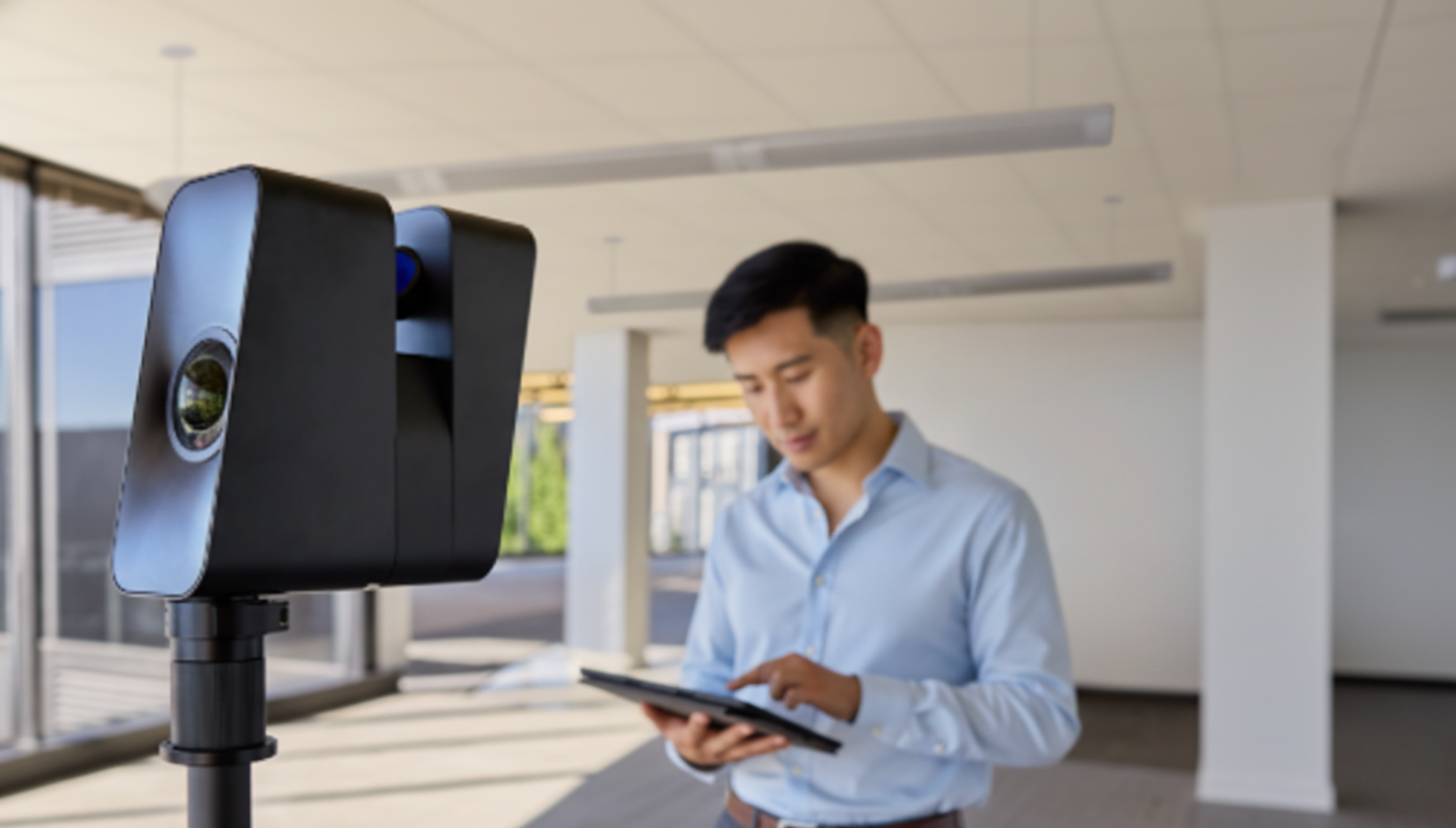 Young man in a light blue shirt using a tablet near a 3D scanning or imaging device indoors with large windows and a modern interior.