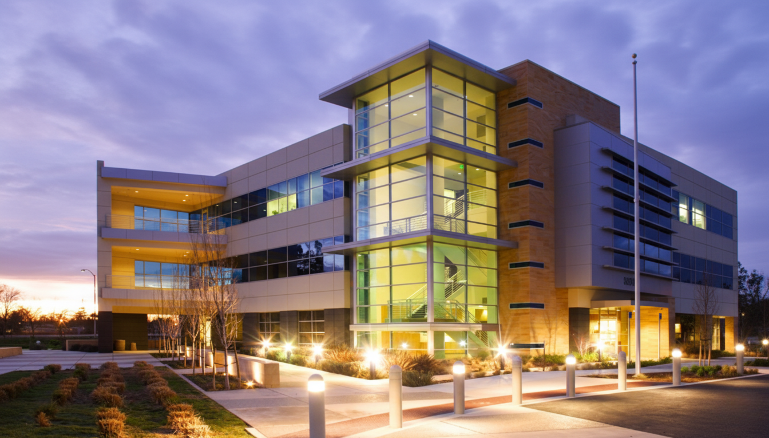 Modern multi-story office building with large glass windows, illuminated from within, during dusk. The building features a prominent glass staircase and landscaped exterior with pathway lights, trees, and a flagpole in the foreground.
