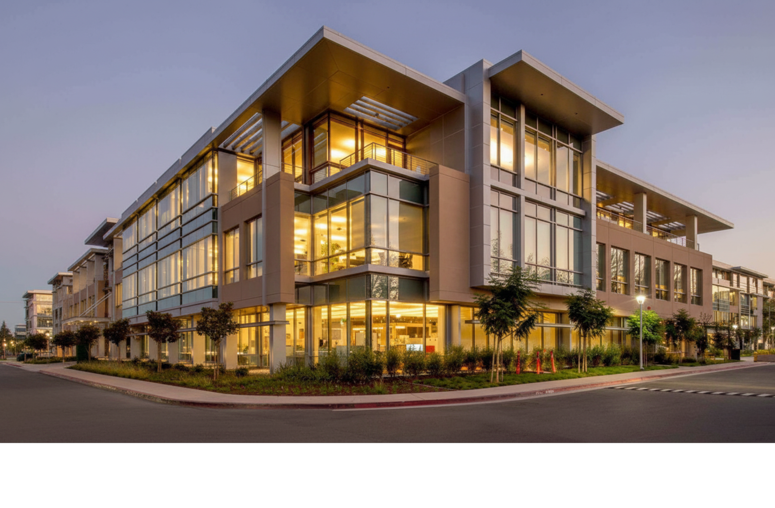 Modern multi-story office building with glass exterior, illuminated interior, and landscaped surroundings at dusk.