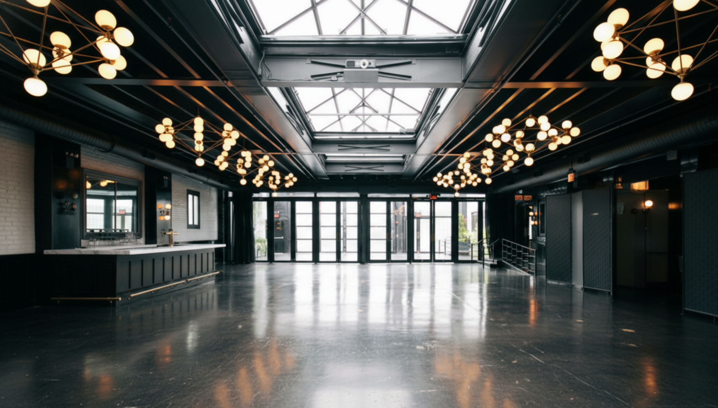 Empty modern event space or lounge with black walls and ceiling, multiple chandeliers with round bulbs, large glass entrance doors, and skylights.