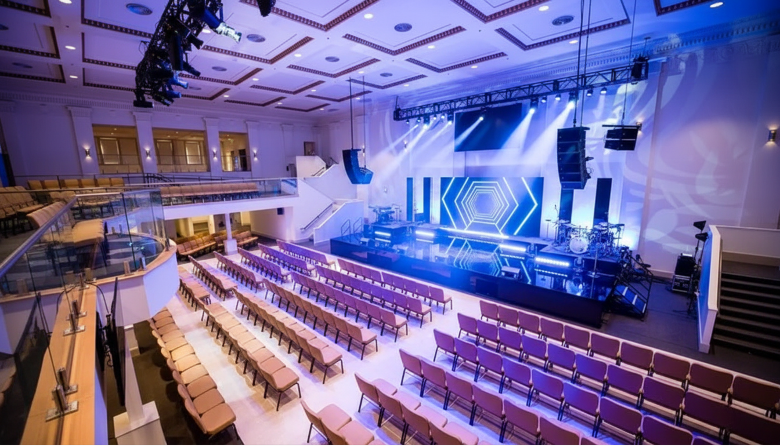 Empty auditorium with a stage, arranged with rows of pink and beige chairs, and a bright LED screen with geometric patterns, stage lights, musical instruments, and speaker systems.
