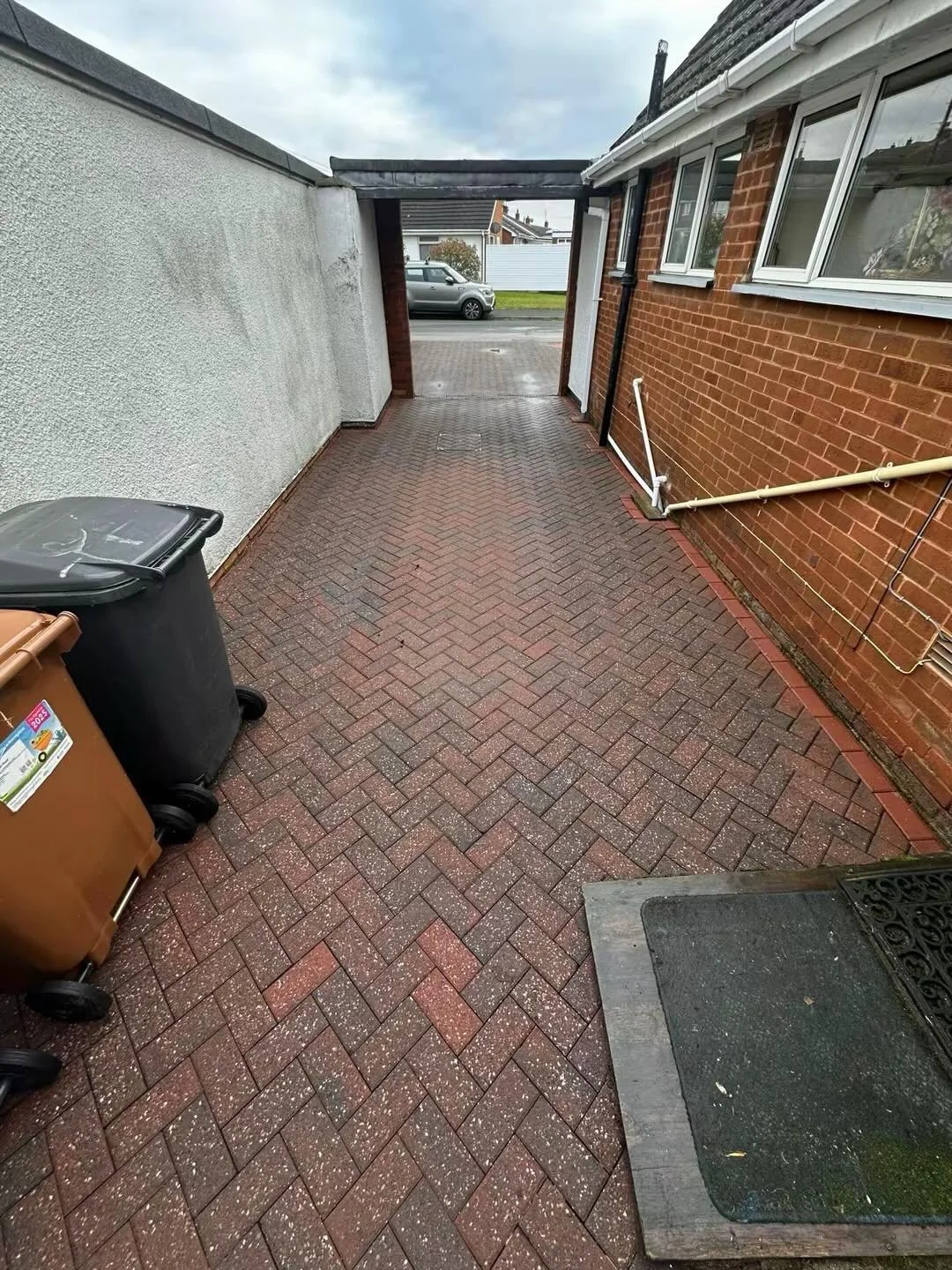 A narrow brick-paved outdoor walkway between a white textured wall and a red brick house. Two trash bins are on the left, and a door mat in front of a door on the right. Piping and drain pipes are attached to the brick wall, with an open gate at the end facing the street with a parked car.