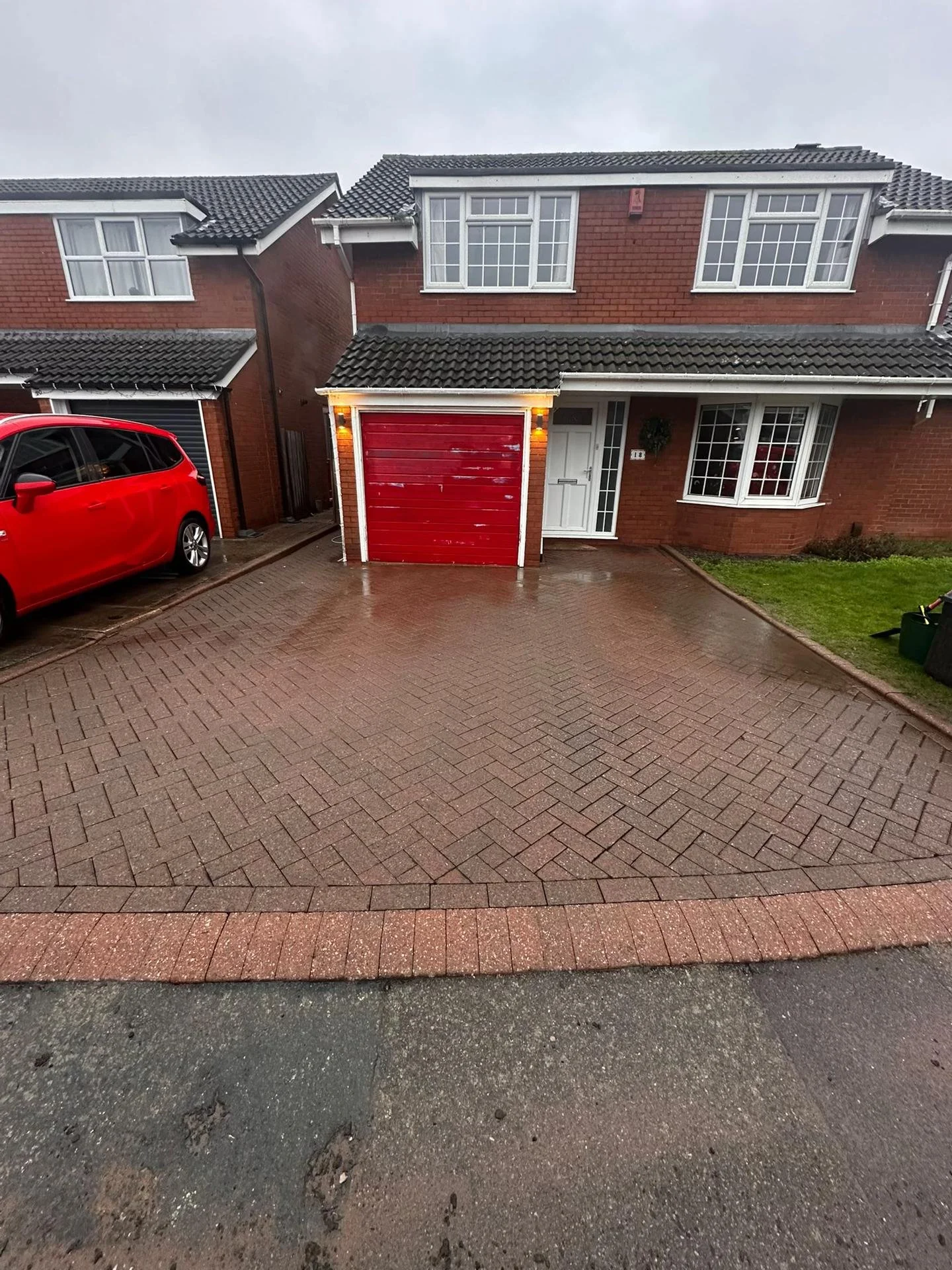 Front of a brick house with a red garage door, a white front door, and a bay window. A small lawn is on the right, with a green trash bin. A red car is parked on the driveway on the left. The driveway and sidewalk are wet, indicating recent rain.