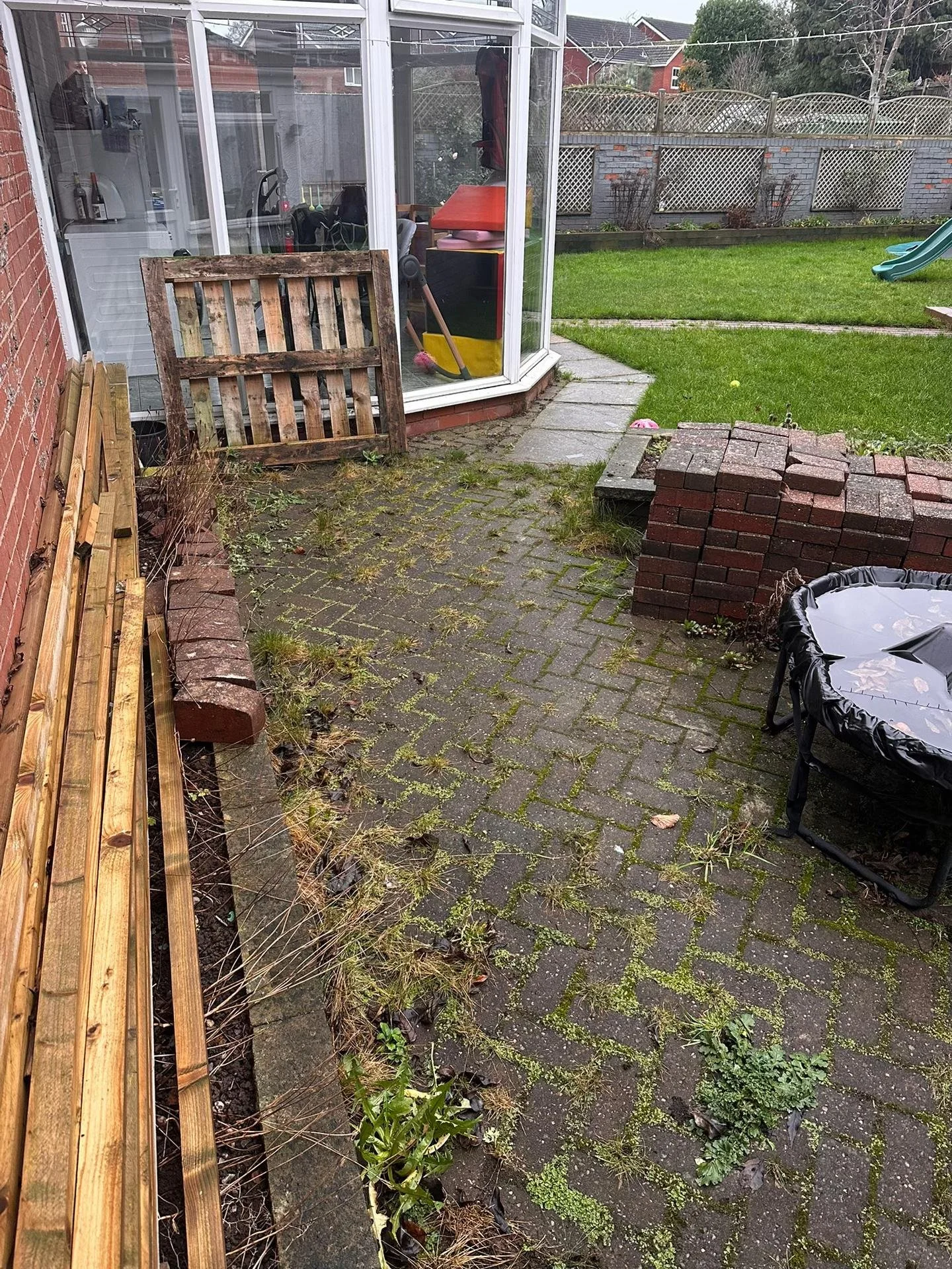 Backyard patio with brick pavement, a small circular trampoline, a pile of stacked bricks, a garden bed with weeds, an enclosed porch with gardening tools, and a grassy area with a slide in the background.