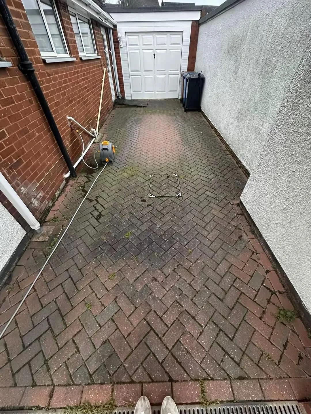A small enclosed backyard with brick and white walls, a paved brick floor, a white garage door, a trash bin, and cleaning equipment including a vacuum and a broom. The photo is taken from a standing position, showing the person's shoes at the bottom of the frame.