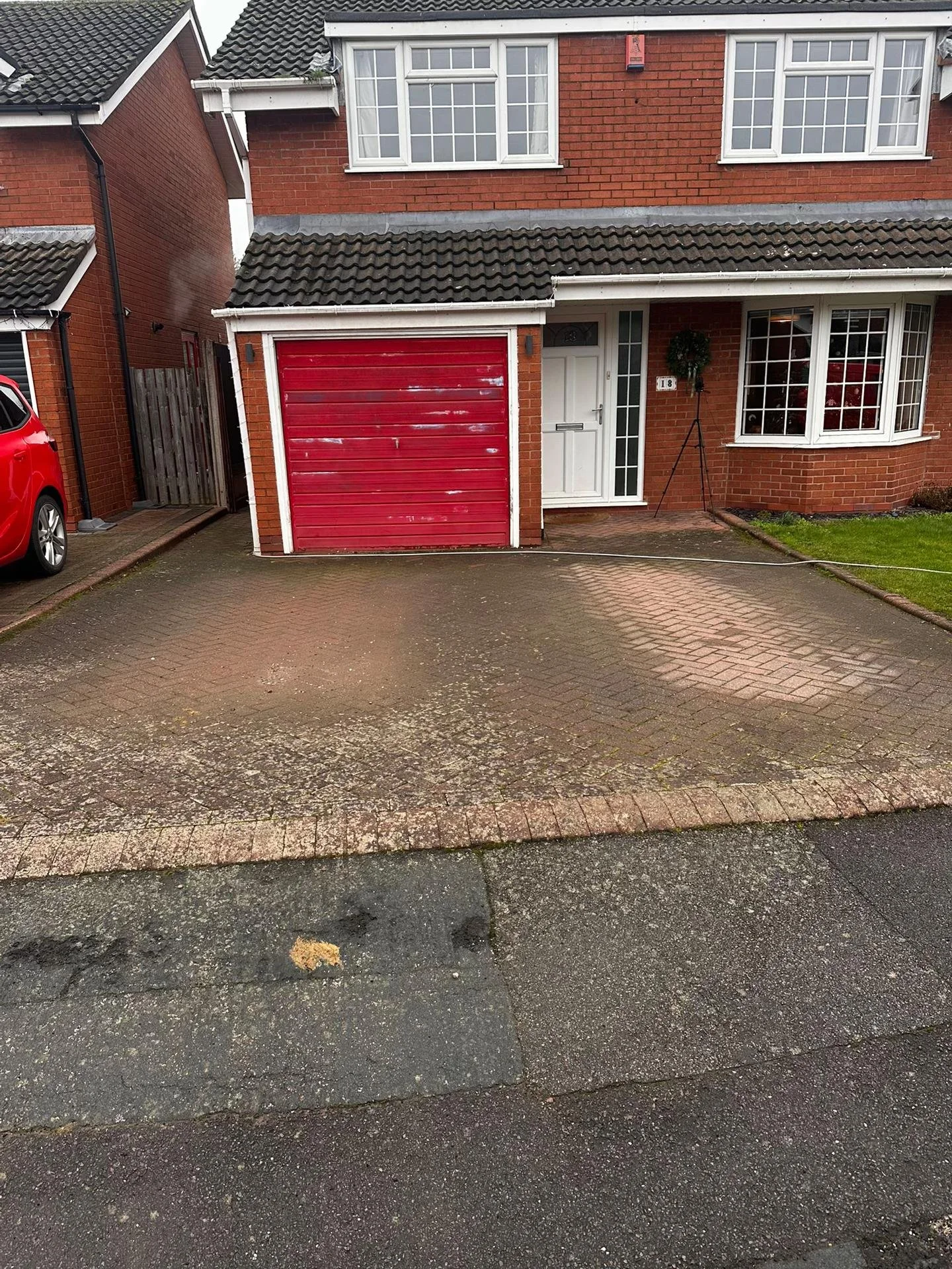 Front view of a brick house with a red garage door, white front door, and a large bay window with a wreath, with a driveway and parked car visible.