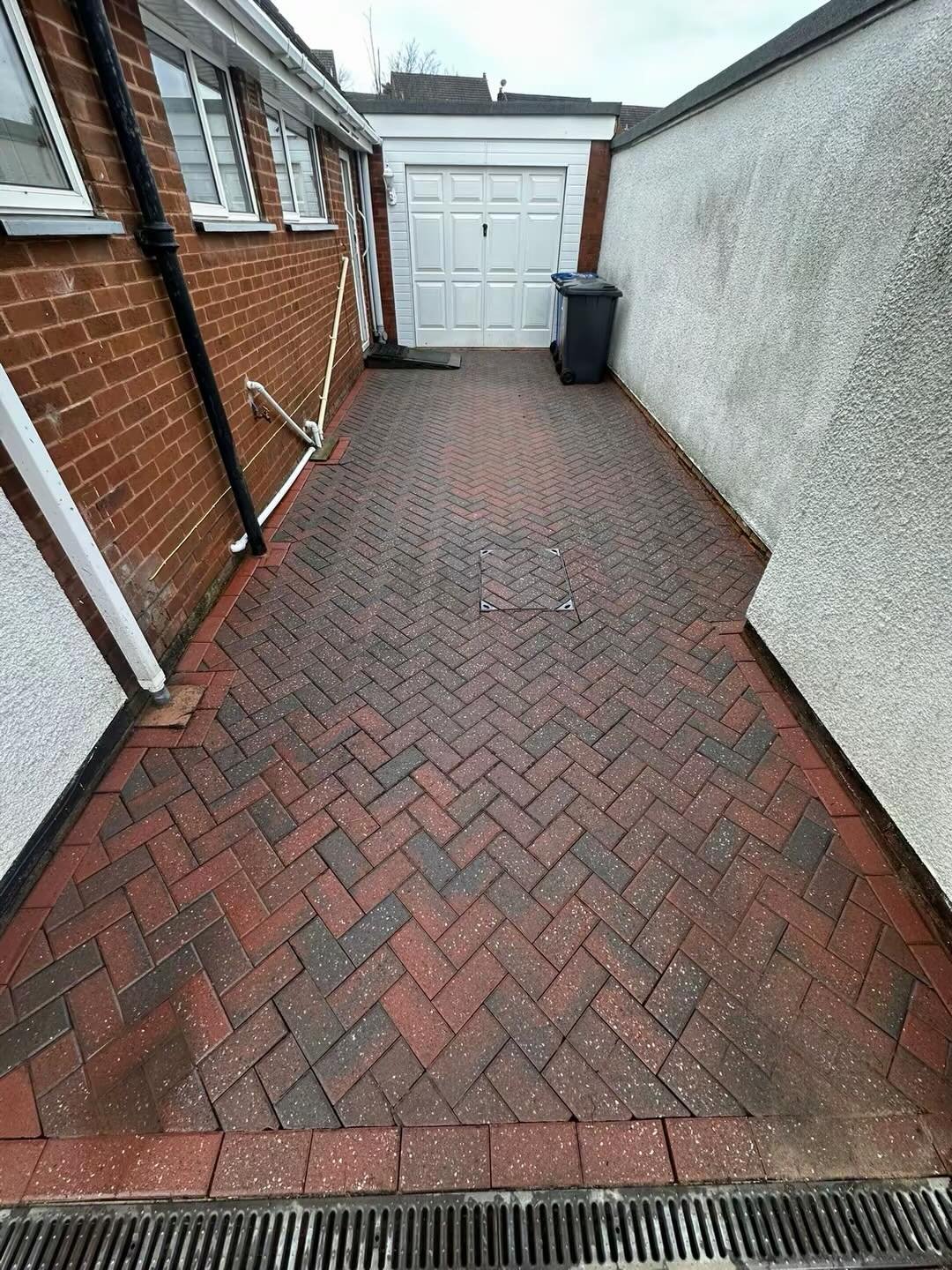 A brick-paved outdoor alleyway between a brick house and a white wall, with a white garage door at the end, black trash bins, and a drain grate at the foreground.