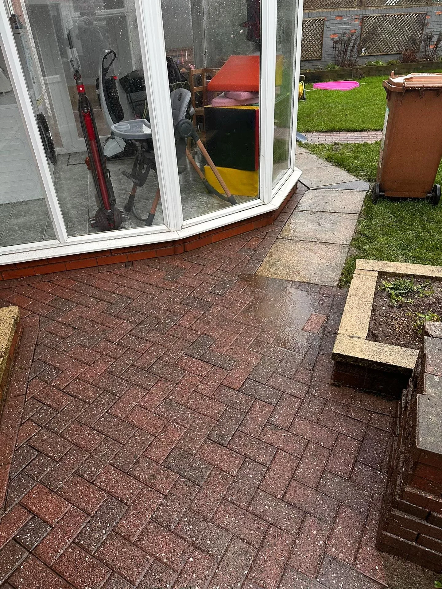View of a backyard with a brick patio, a glass enclosed sunroom, a brown trash bin, and some garden beds. The sunroom contains exercise equipment and a small table with chairs inside.