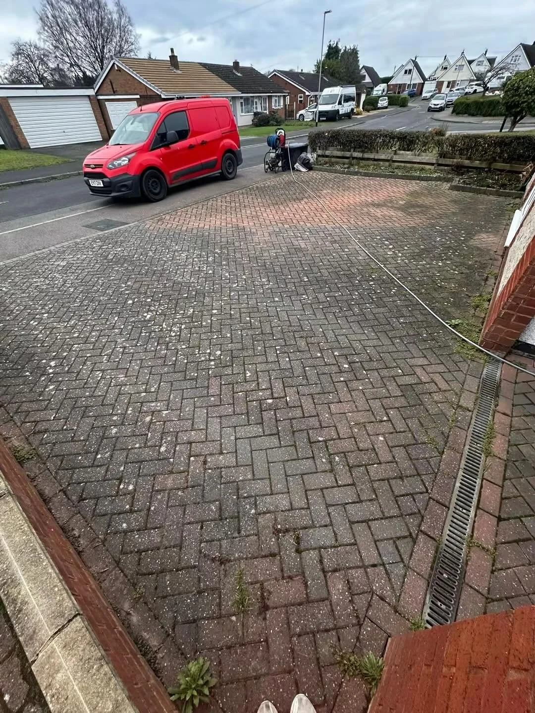 View of a brick driveway with a red van parked on the street, a gardening trolley, and a garden hose reel, with houses and cars in the background under a cloudy sky.