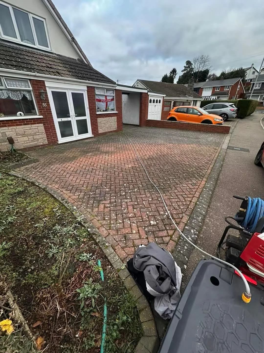 A residential house with a brick driveway, parked cars, and garden area, on a cloudy day.