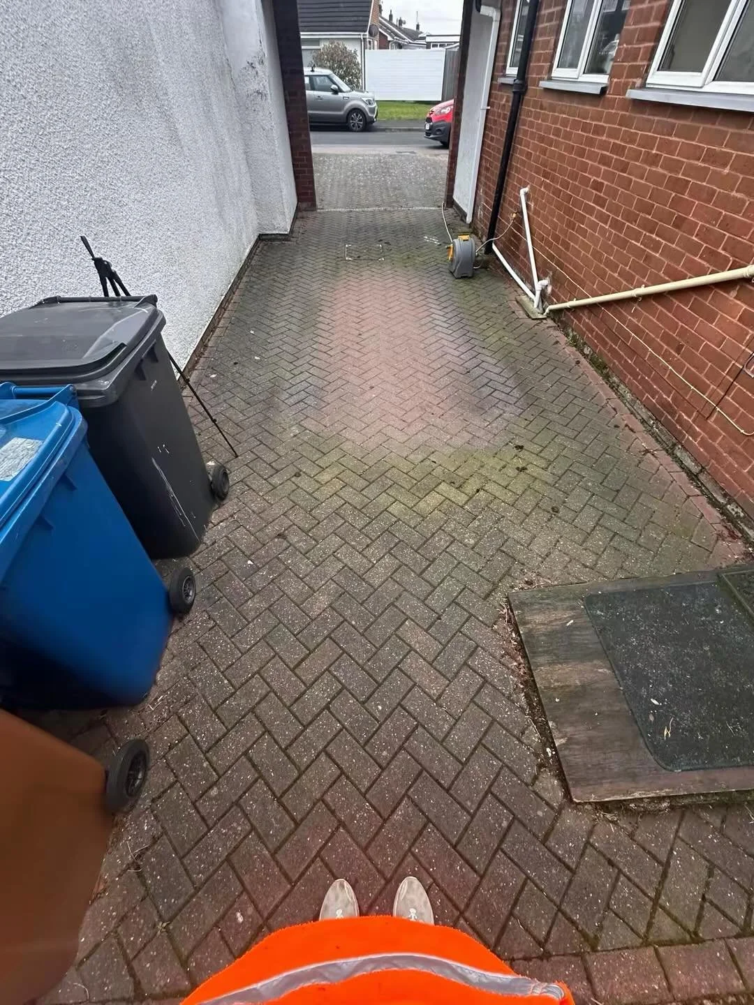 A narrow paved alleyway between a white and a red brick building, with trash bins on the left, a garden hose reel on the right, and a glimpse of parked cars on the street beyond.