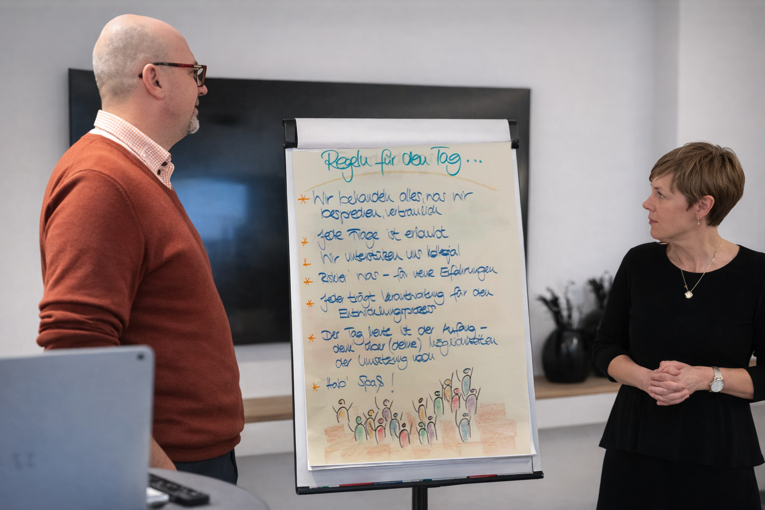 Zwei Menschen in einem Büro, die über ein Whiteboard mit Regeln für den Tag sprechen.