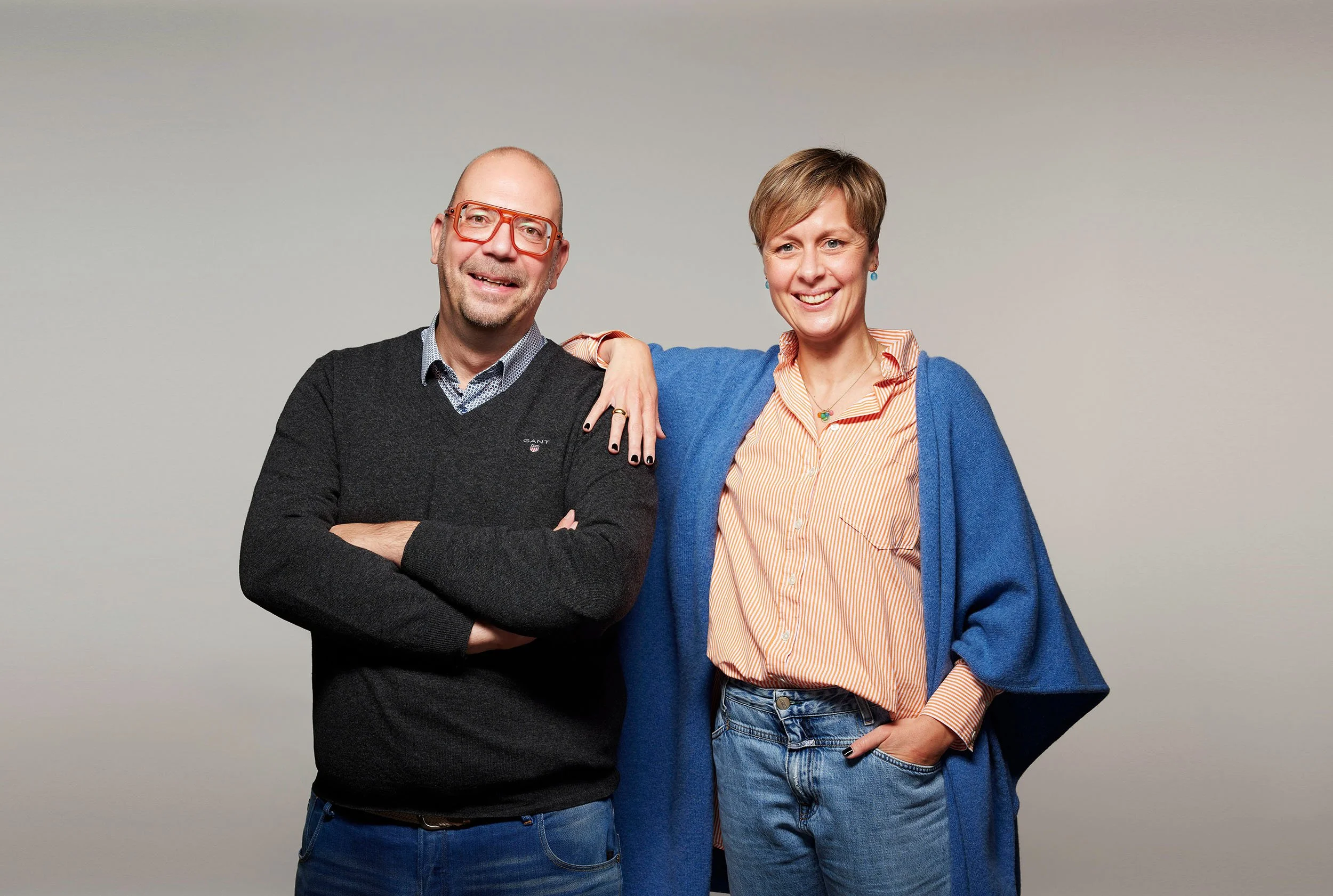 Ein Mann und eine Frau stehen lächelnd vor einem neutralen Hintergrund. Der Mann trägt eine schwarze Pullover und rote Brillen, die Frau trägt ein gestreiftes Hemd, eine blaue Jacke und Jeans.