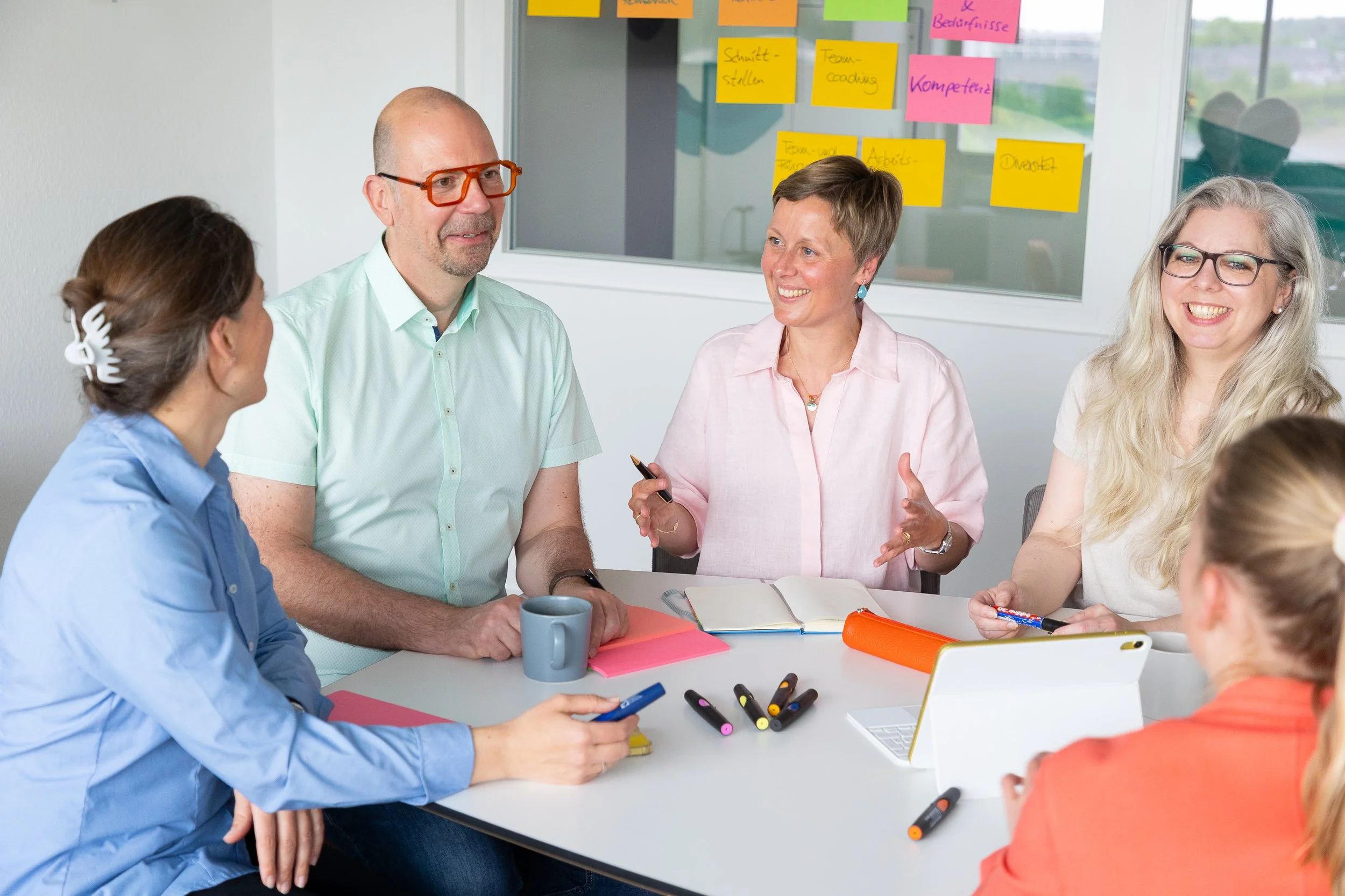 Fünf Personen sitzen an einem Tisch in einem Meetingraum, diskutieren und lachen. Im Hintergrund sind farbige Haftnotizen an der Wand.