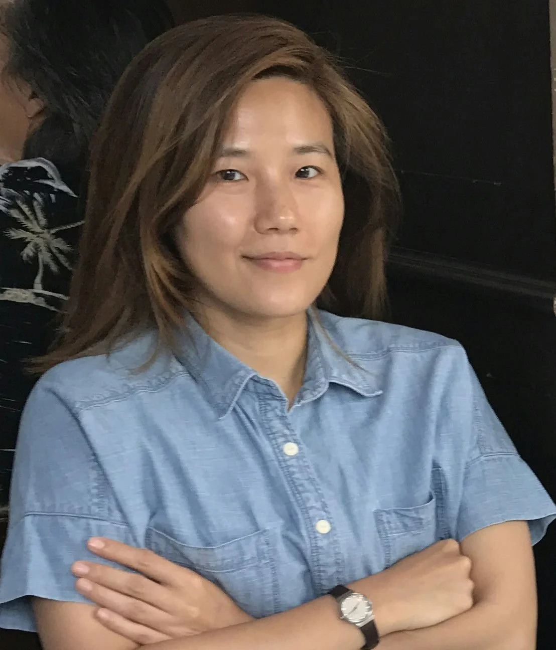 A woman with shoulder-length brown hair, wearing a light blue denim shirt, sitting with arms crossed and smiling slightly.