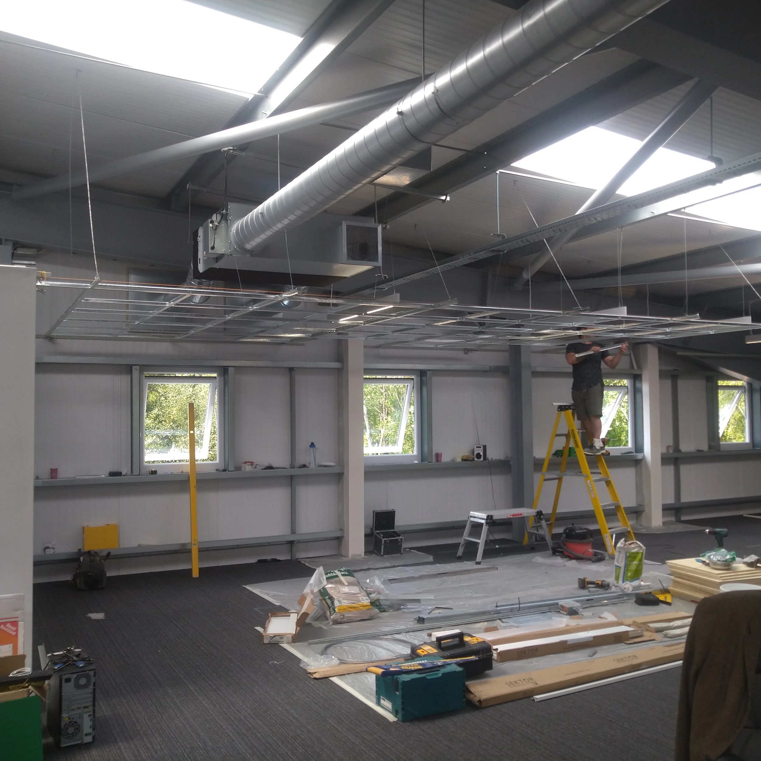 A worker installing ceiling fixtures in an industrial or commercial space with high windows, tiled flooring, and visible ductwork.