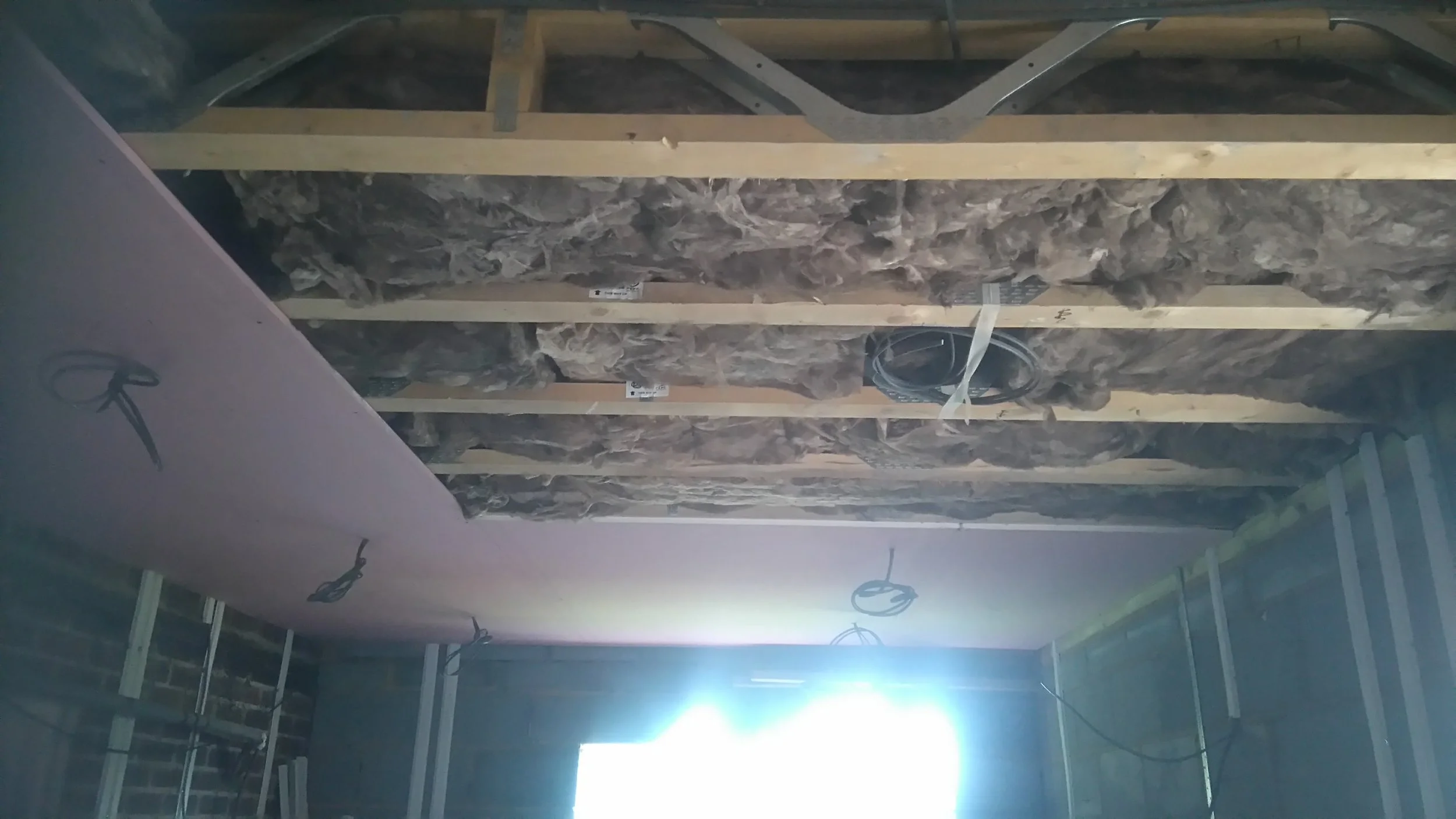 Ceiling construction with exposed wooden beams, insulation material, and electrical wiring, with a window allowing natural light into the room.